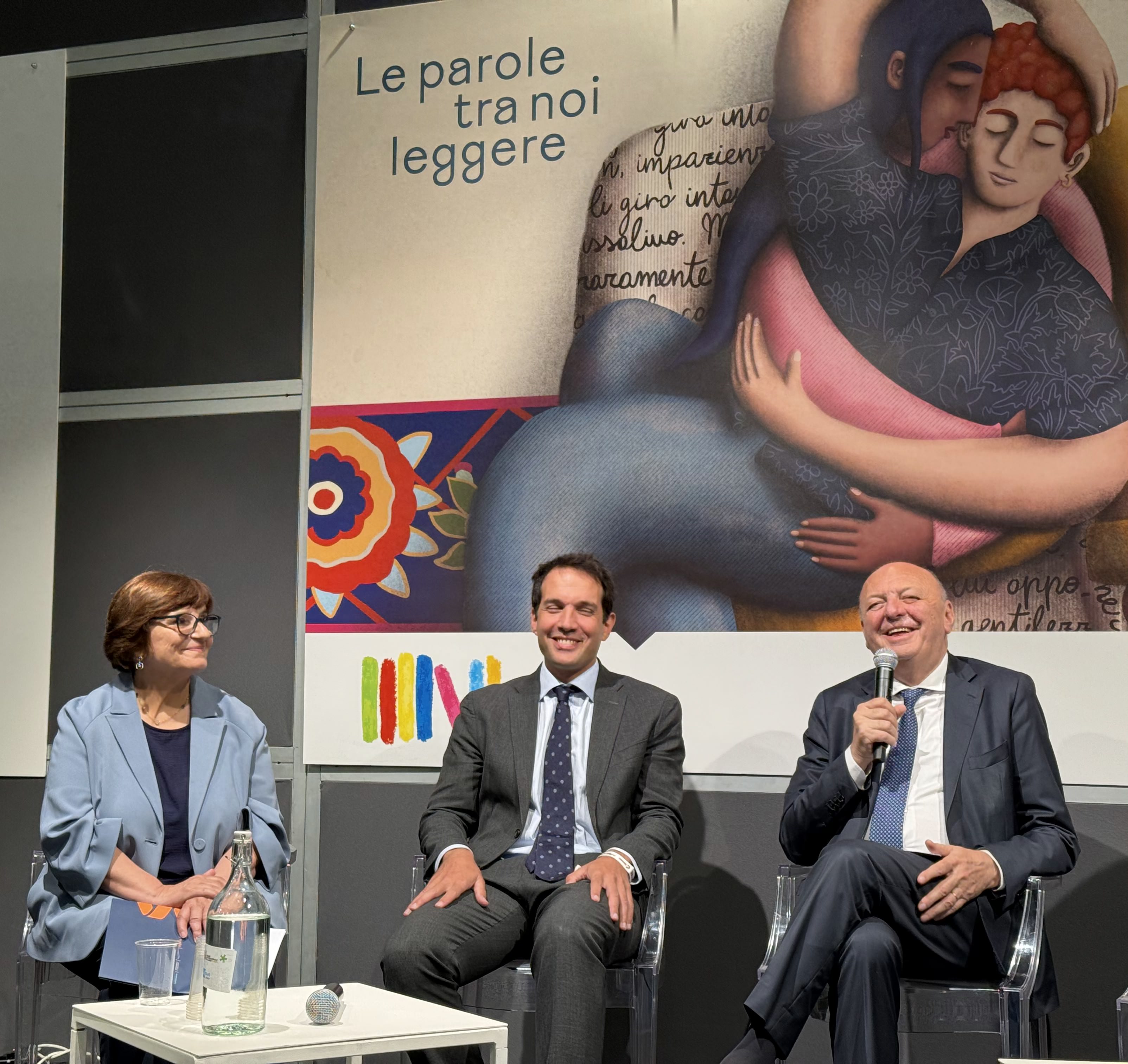 Three people seated on stage, engaged in conversation, with an art backdrop featuring two figures embracing.
