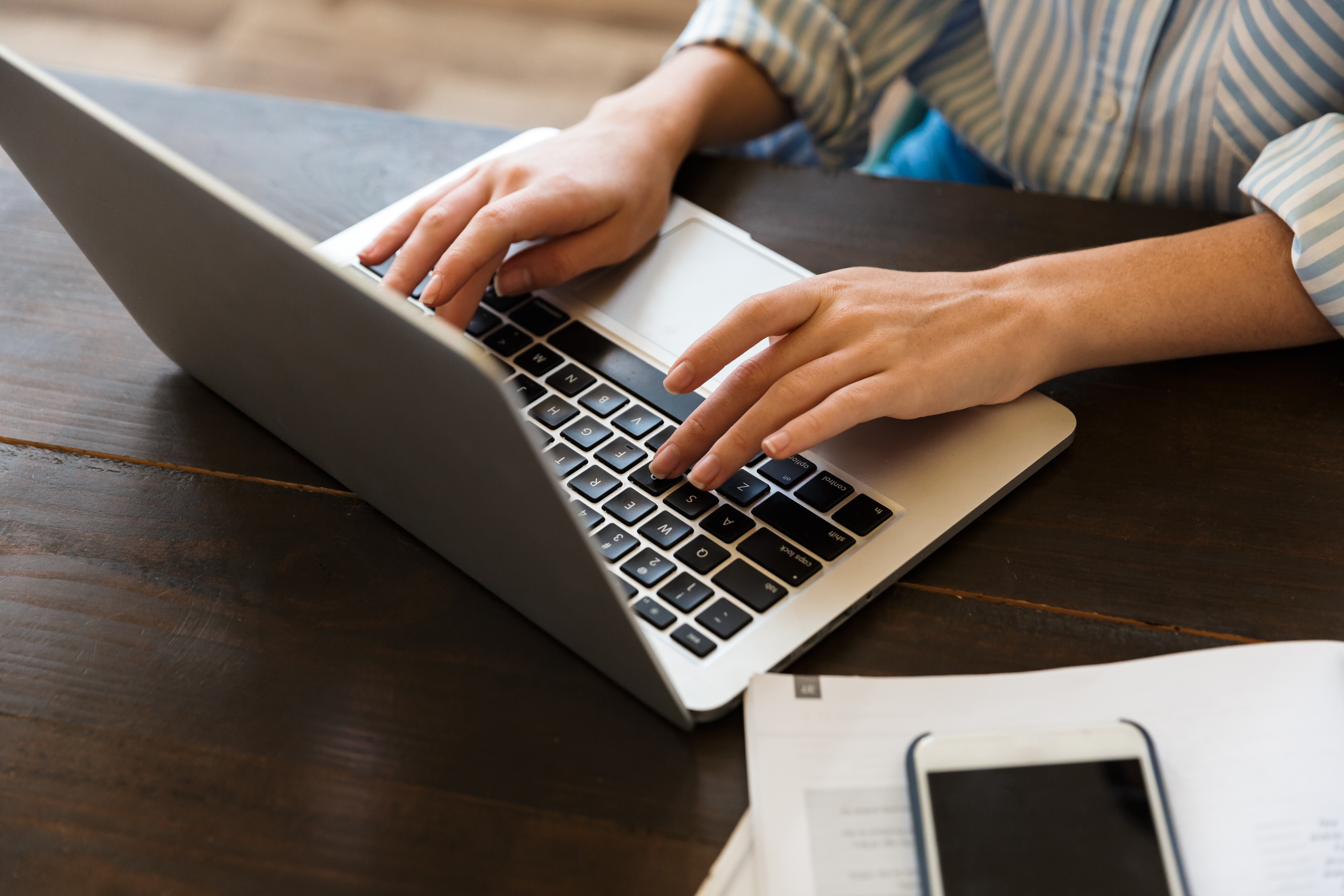 Hands typing on a laptop keyboard beside a smartphone and documents on a wooden table.