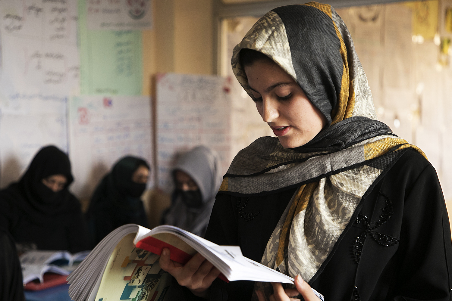 A young girl in a headscarf reads a book, with classmates in the background.