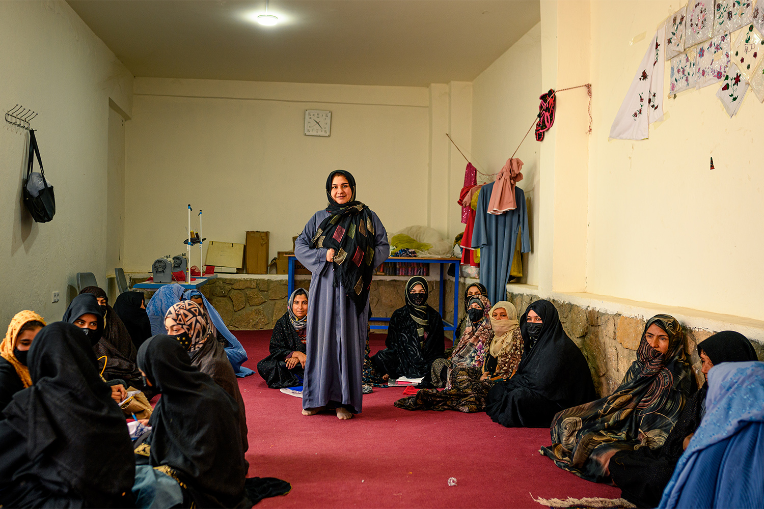 Women in traditional attire gather in a room, seated on the floor, with one woman standing.