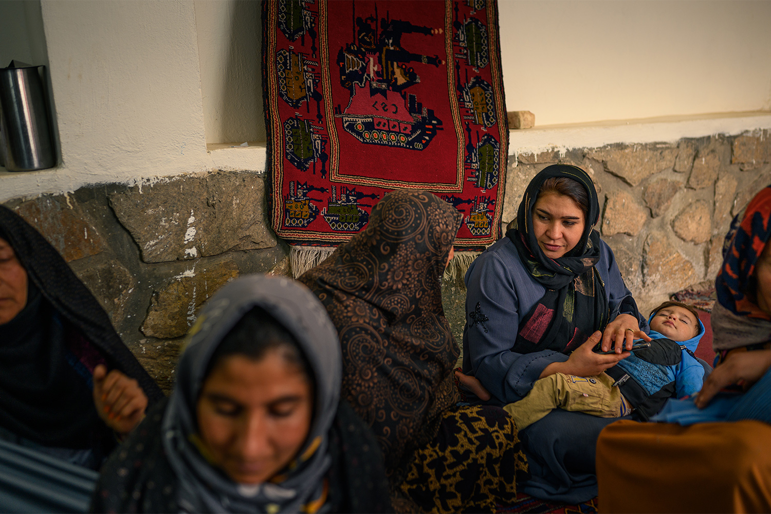 Several women in traditional attire sit together, one holding a baby, in a cozy indoor setting.