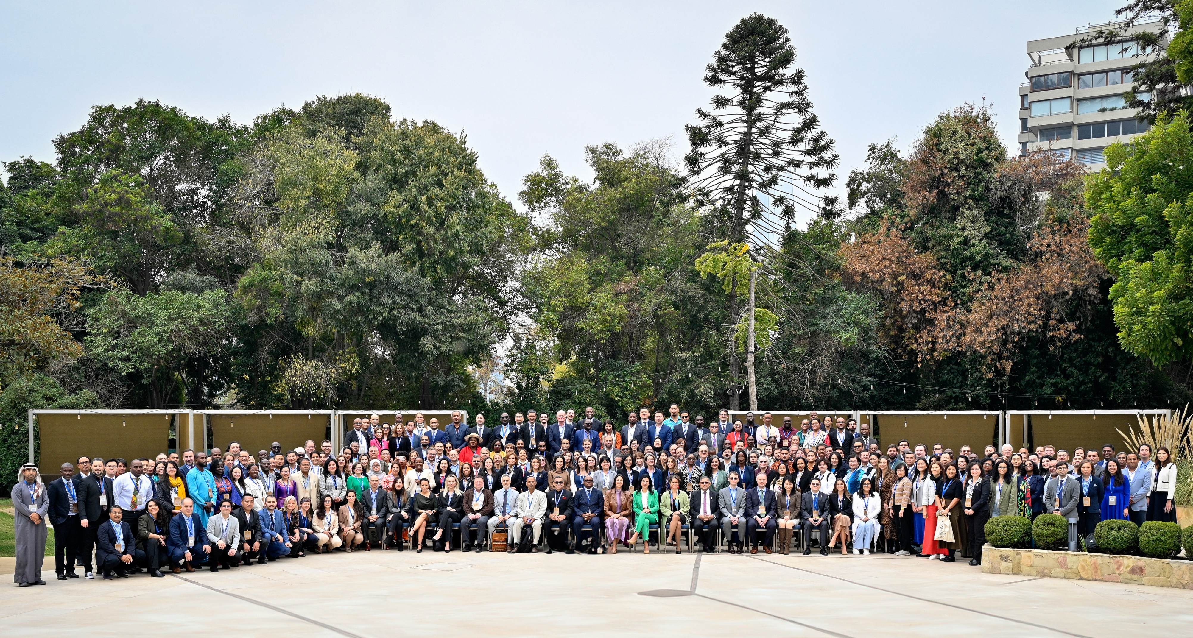 A large group of people gathered outdoors, standing in rows with greenery in the background.