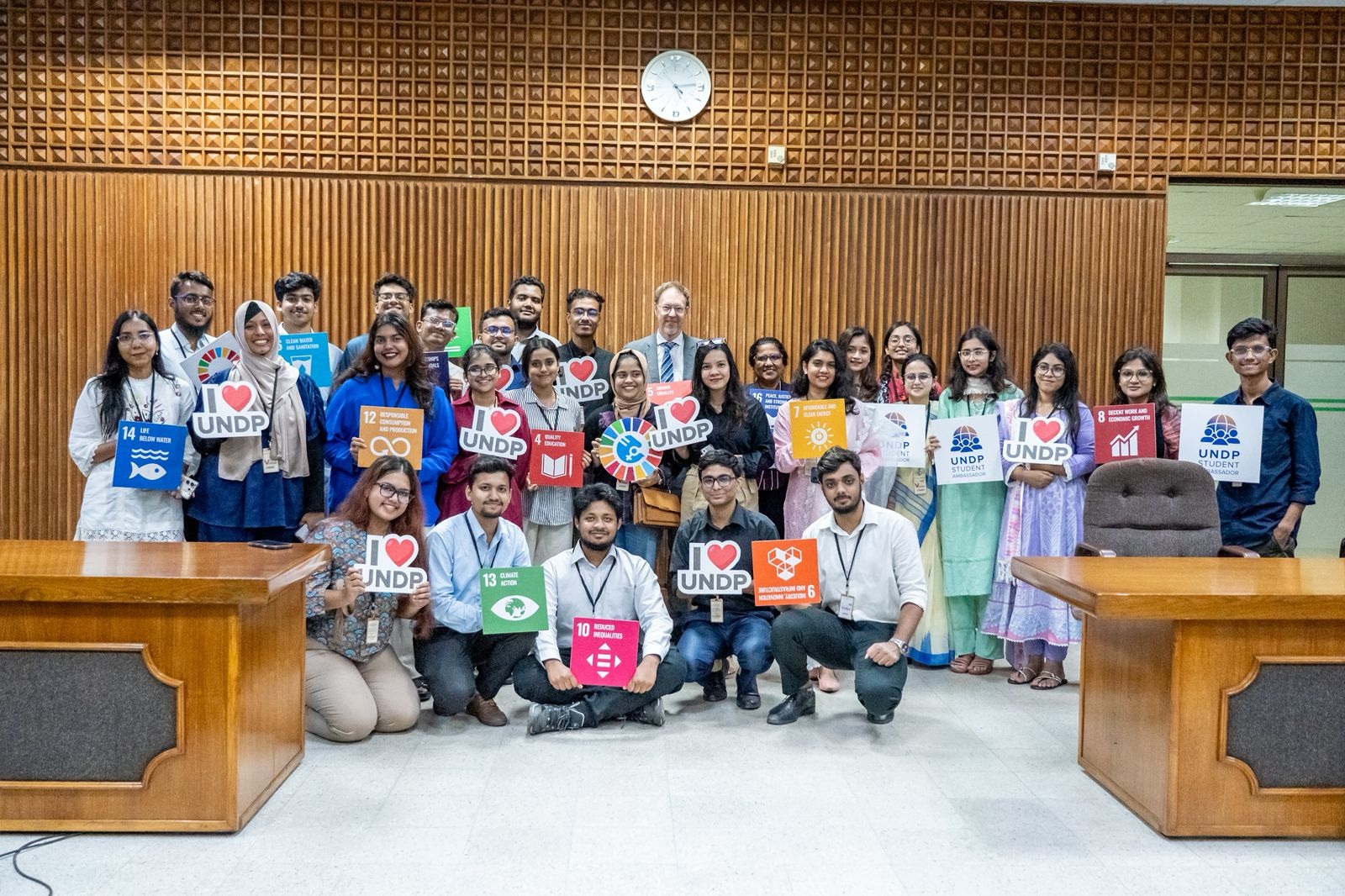 Group photo of diverse individuals holding colorful signs, smiling in a conference room.