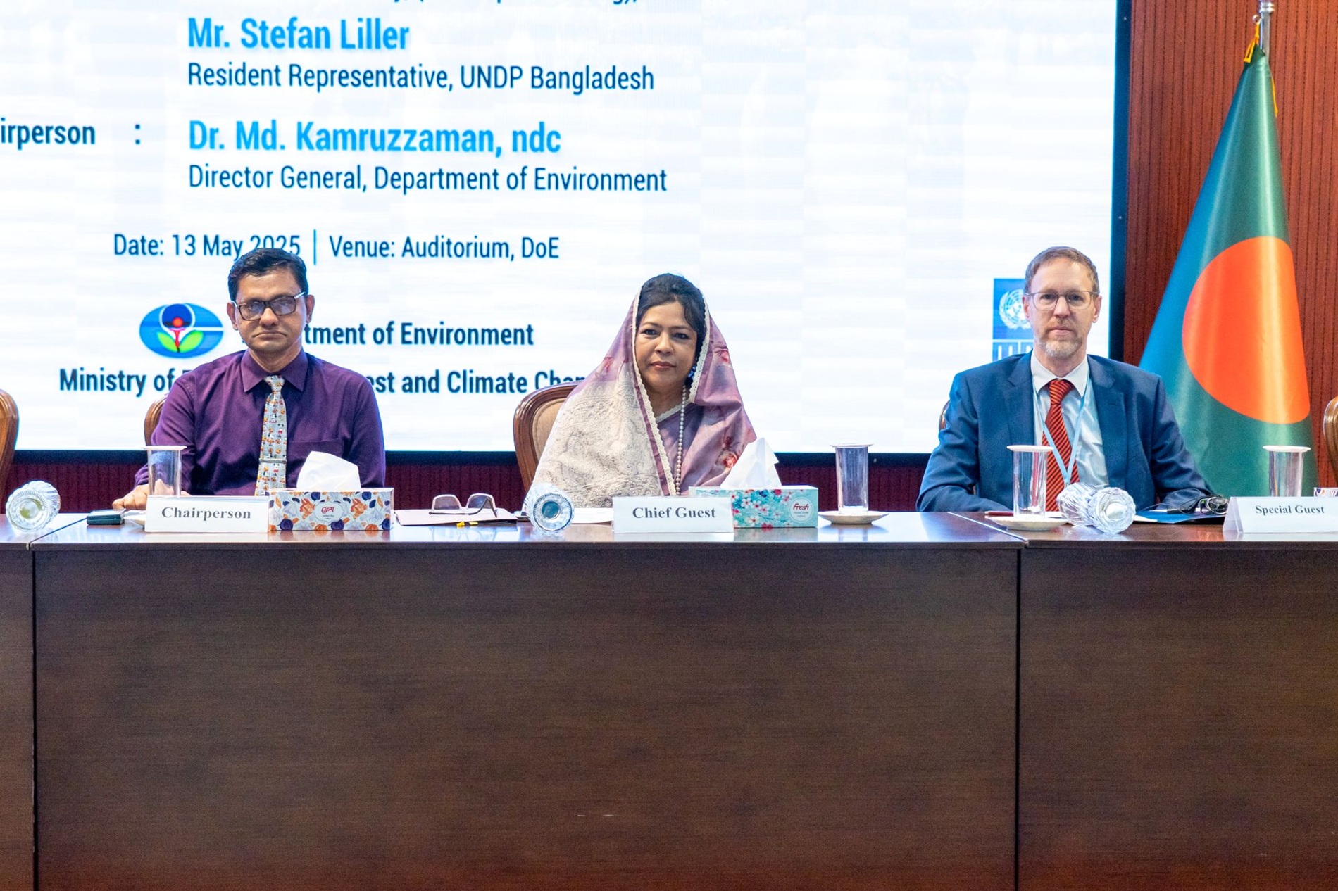 Three seated professionals at a conference table, with a Bangladeshi flag in the background.