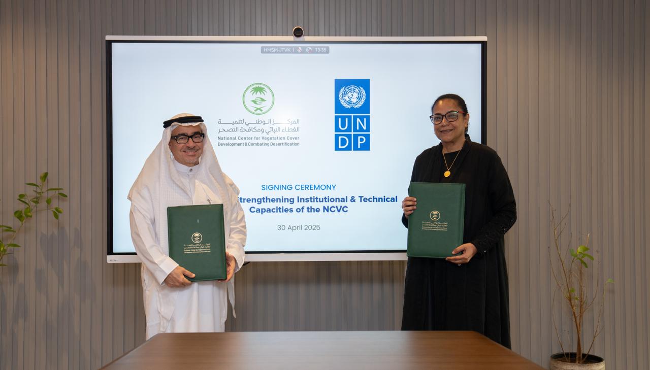 Two individuals in formal attire hold green documents, standing in front of a presentation screen.