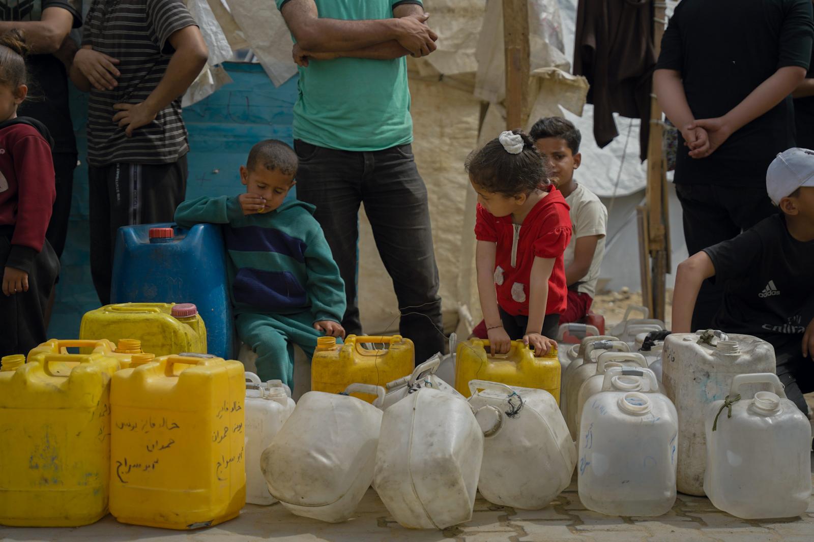 Children and adults stand beside colorful plastic jerrycans in a makeshift camp.