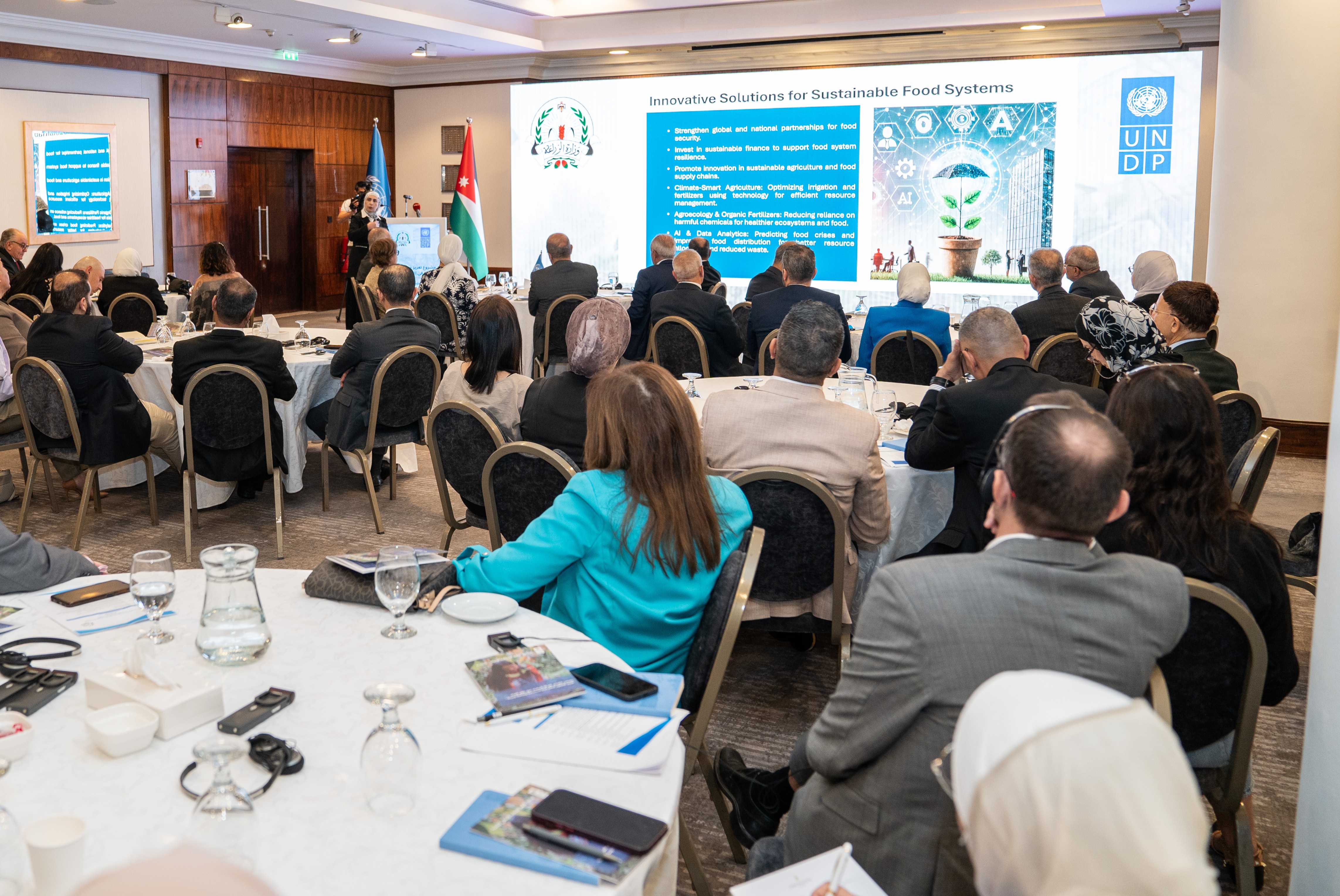 Conference attendees seated at tables, listening to a speaker in front of a presentation screen.