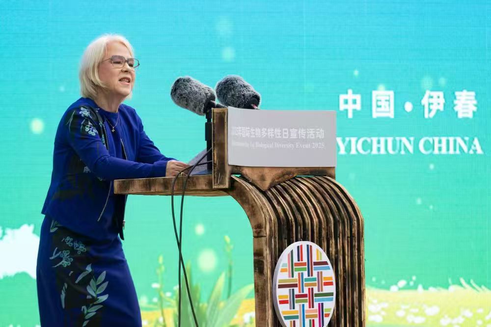 A woman speaking at a podium, with a colorful backdrop featuring flowers and text in Chinese.