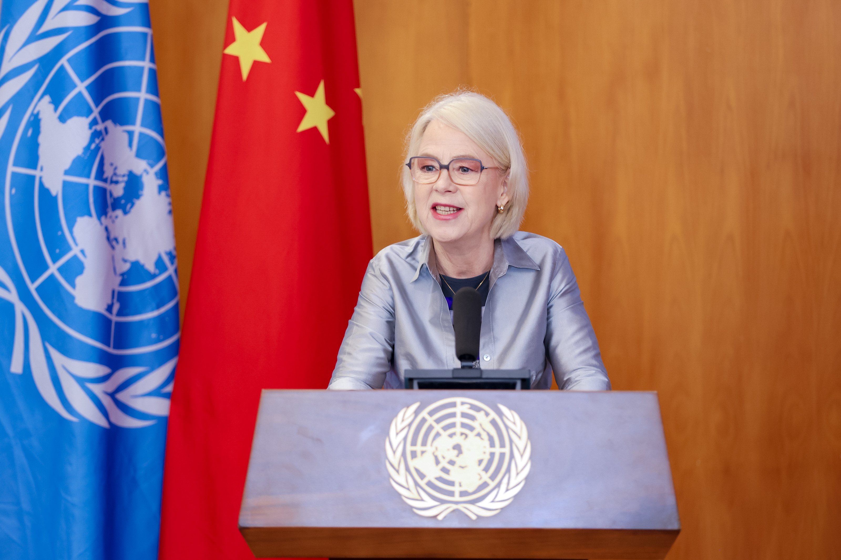 Woman speaking at a podium with UN emblem, flanked by the Chinese flag and a world map backdrop.