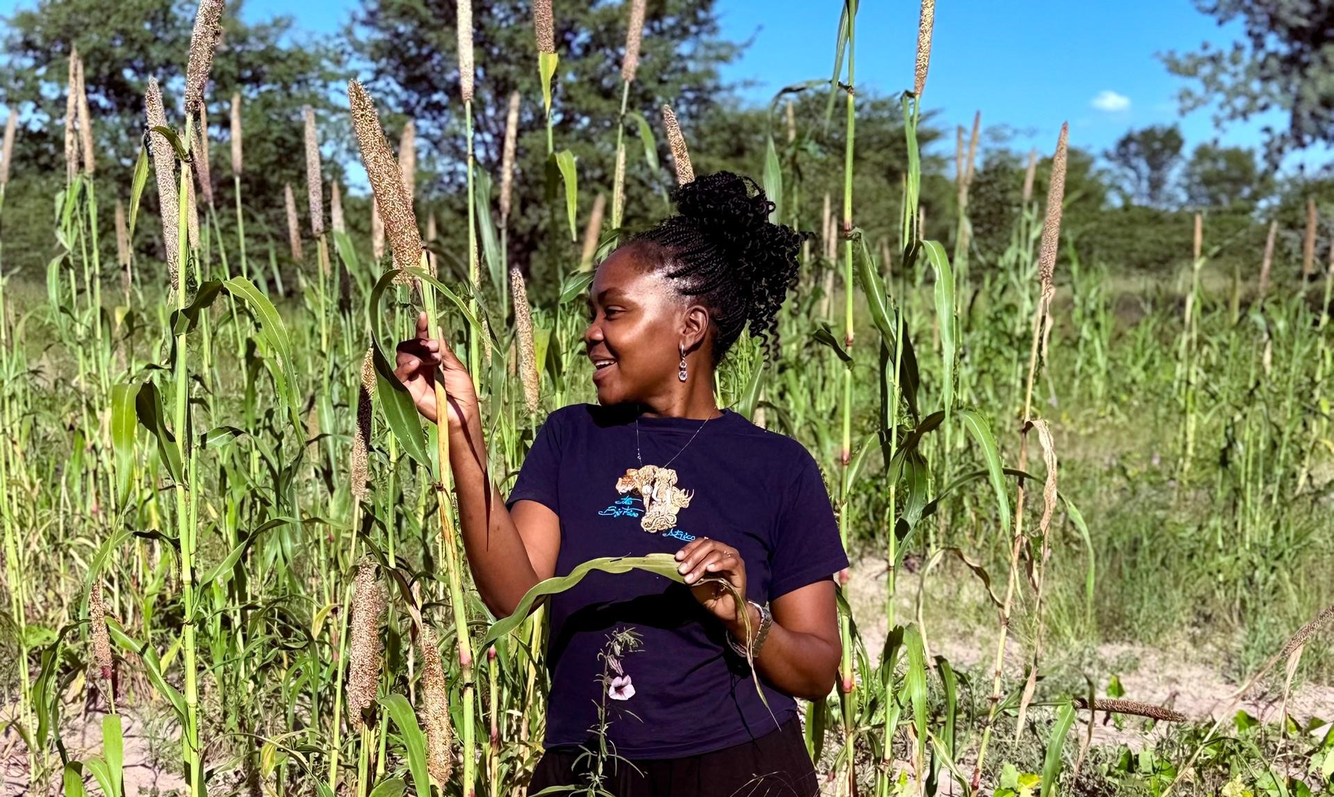 A woman smiles while holding a plant amidst tall green vegetation under a blue sky.