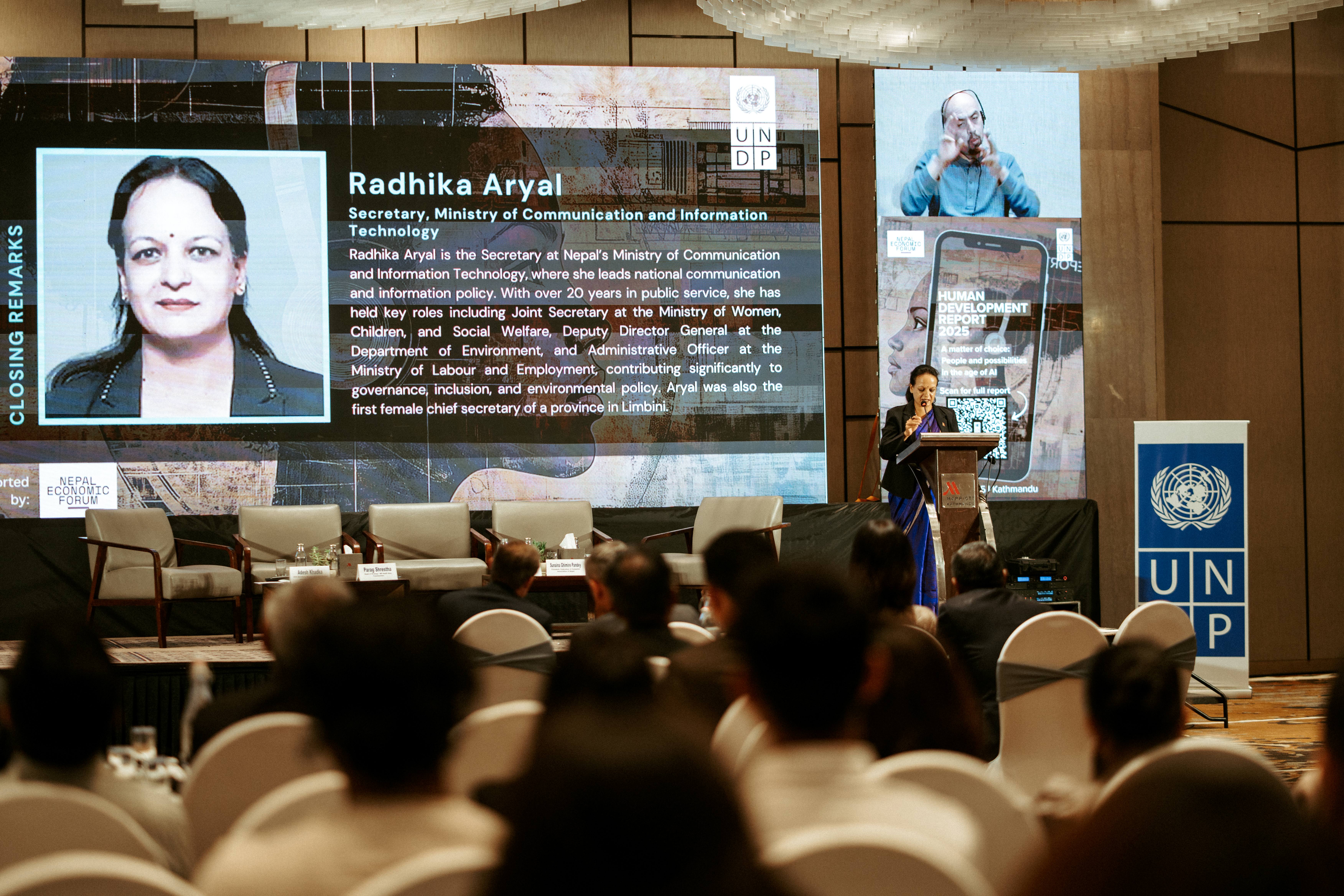 A speaker at a podium addresses an audience, with a large screen displaying their photo and information.