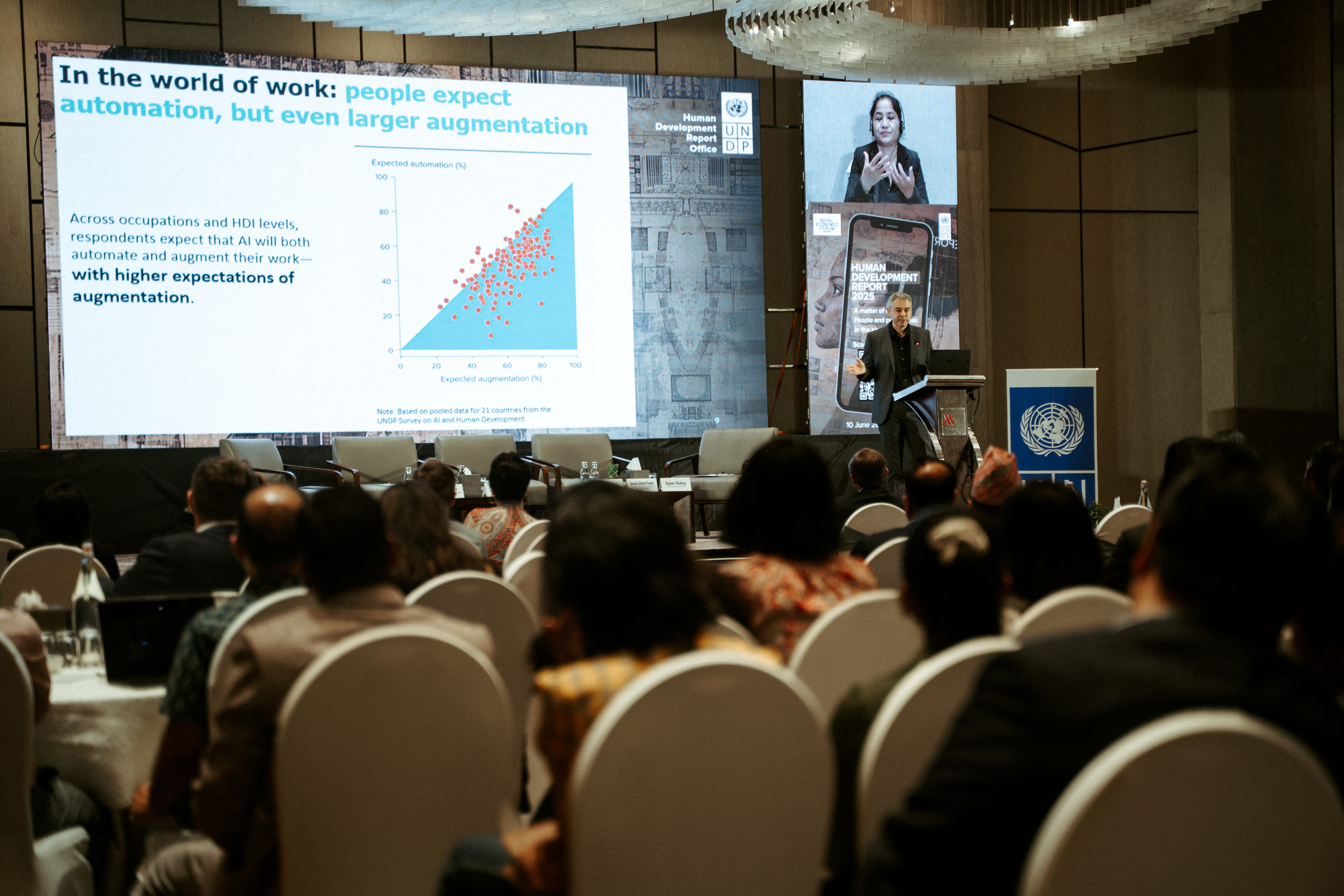 A speaker presents data in a conference hall, with an audience seated and a large screen displaying a graph.
