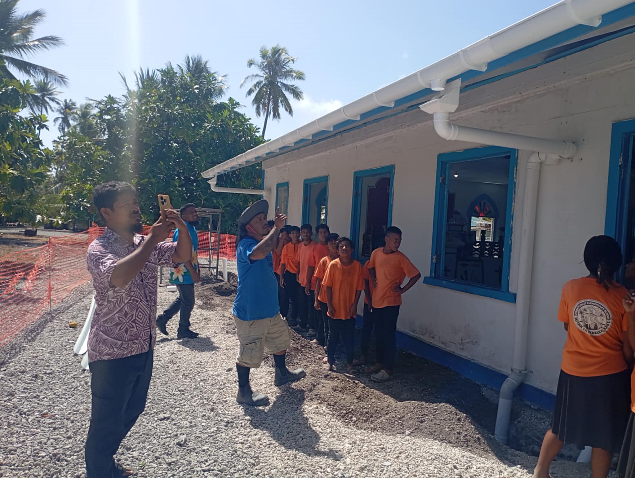 A group of children in orange shirts stands by a building while an adult takes photos.