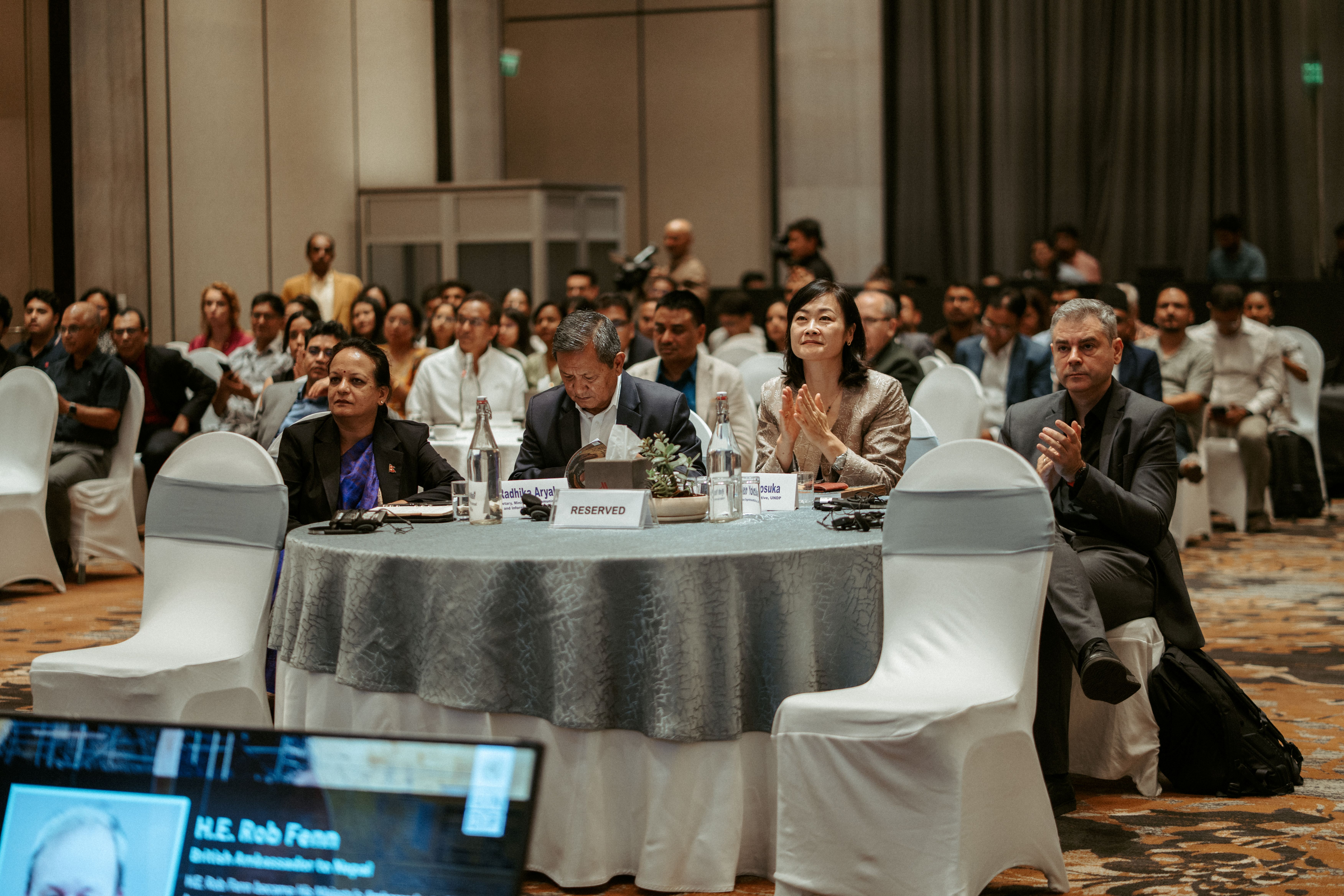 A diverse audience sitting at tables, clapping during a formal event in a spacious hall.