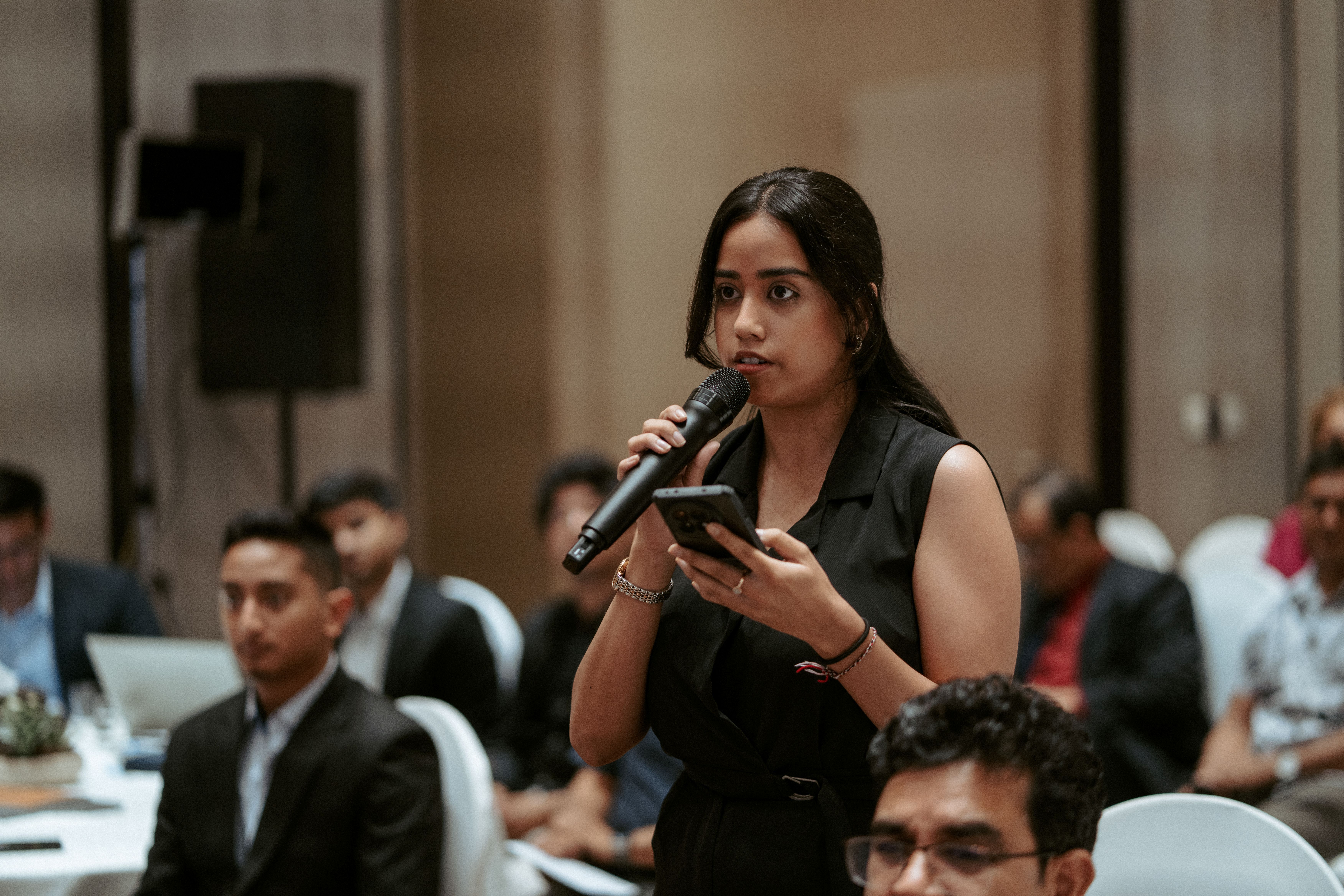 A woman in a black outfit holds a microphone and speaks during a presentation, others seated nearby.