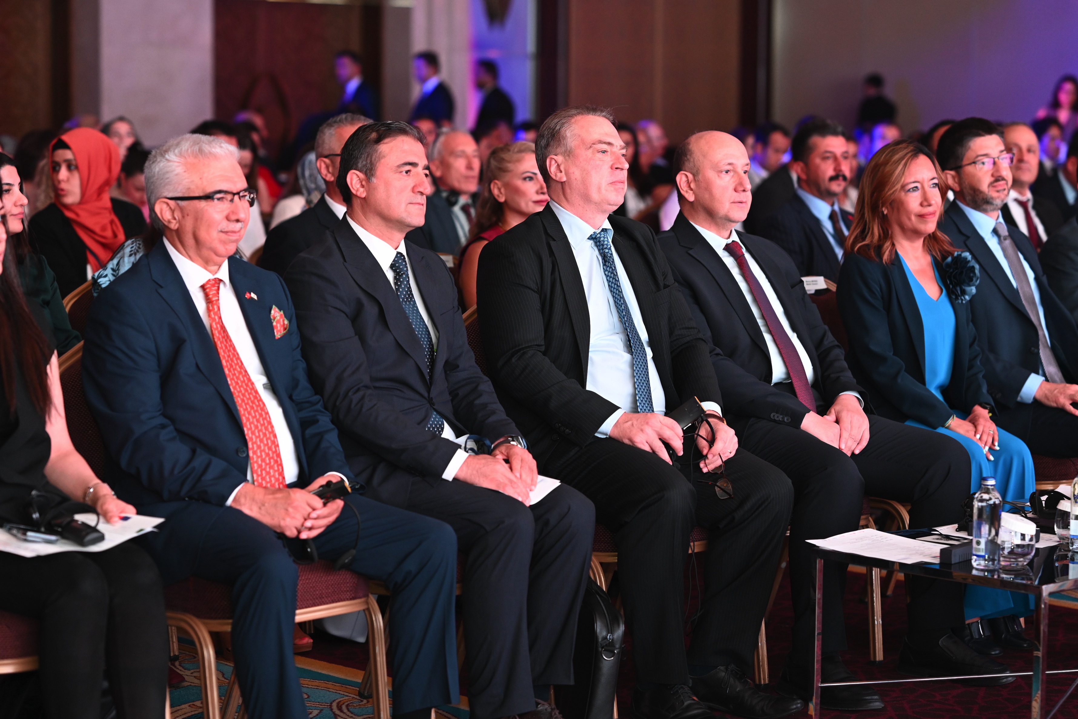 A group of people seated in formal attire at an event, with a diverse audience in the background.