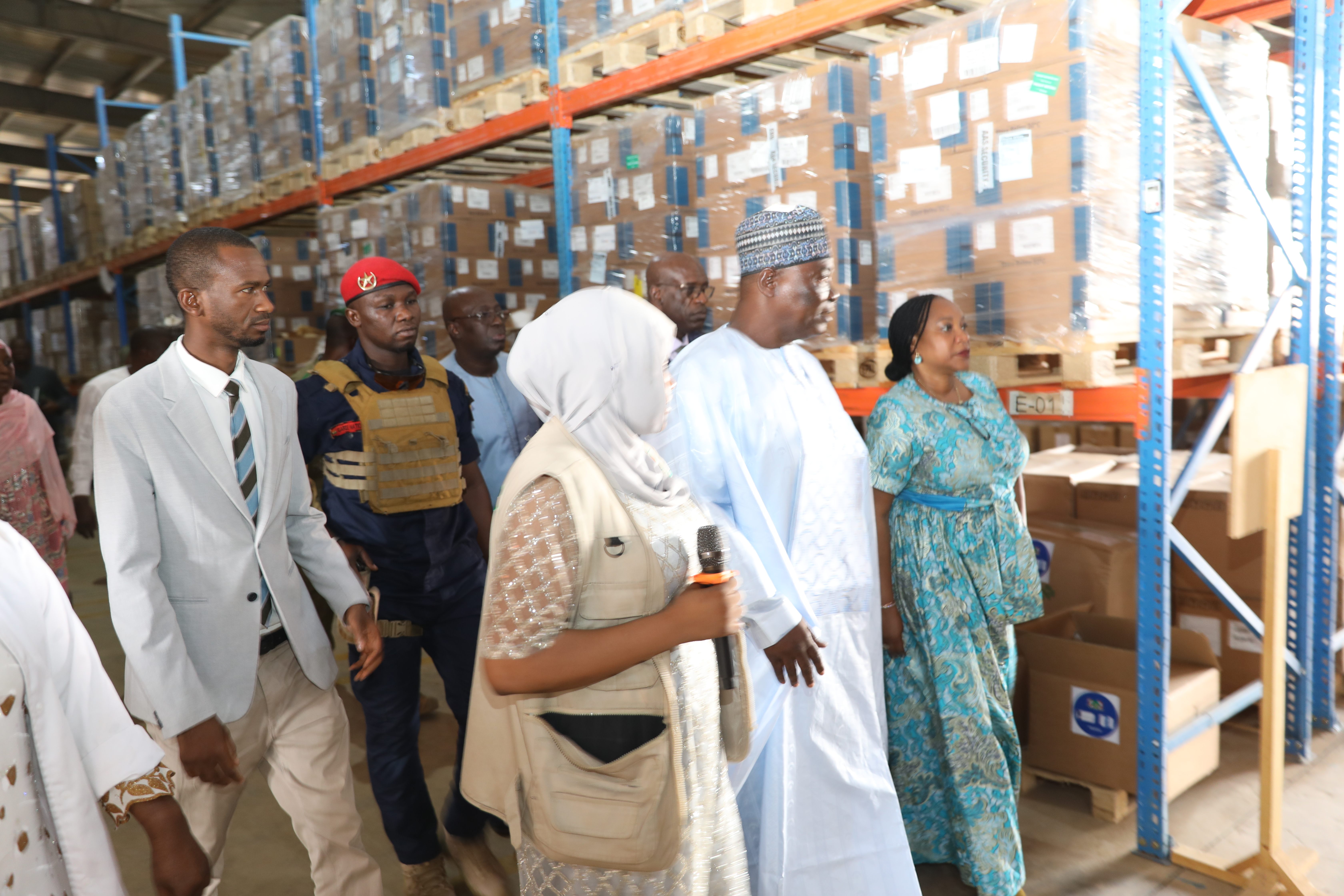 A group of people, including a man in traditional attire, touring a large warehouse filled with boxes.