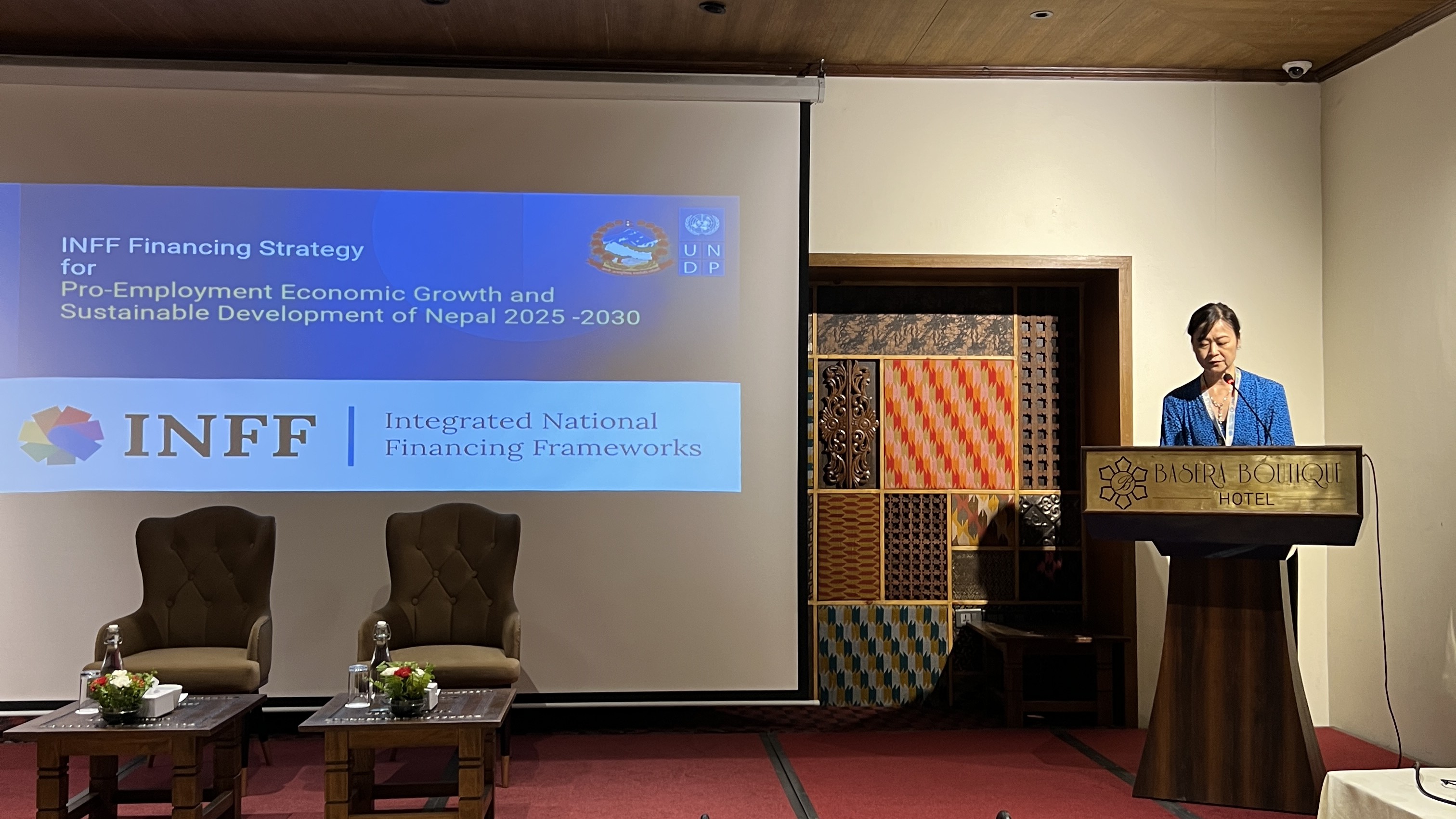 A speaker stands at a podium in a conference setting, with a presentation displayed behind.