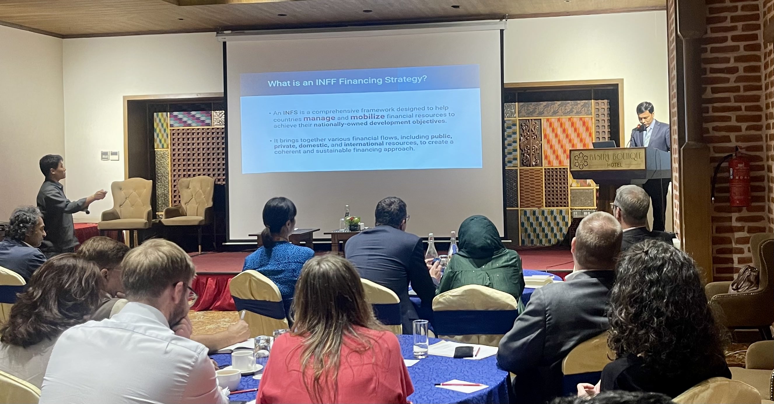 A speaker presents in front of an audience, with a large screen displaying text.