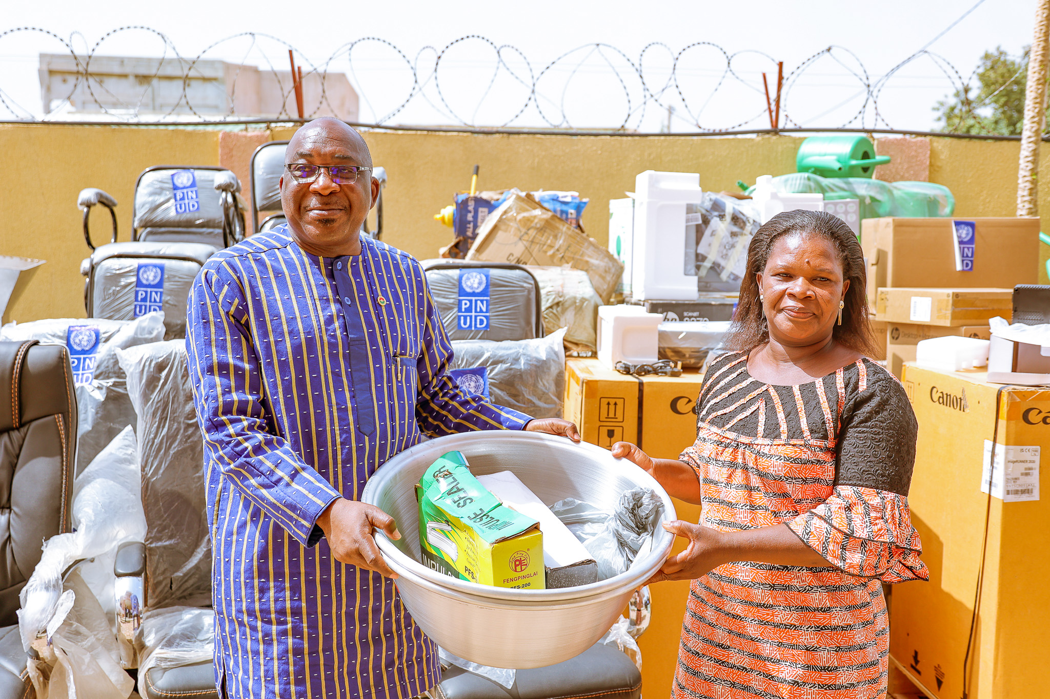 Two smiling individuals are holding a large bowl filled with household items, surrounded by boxes.