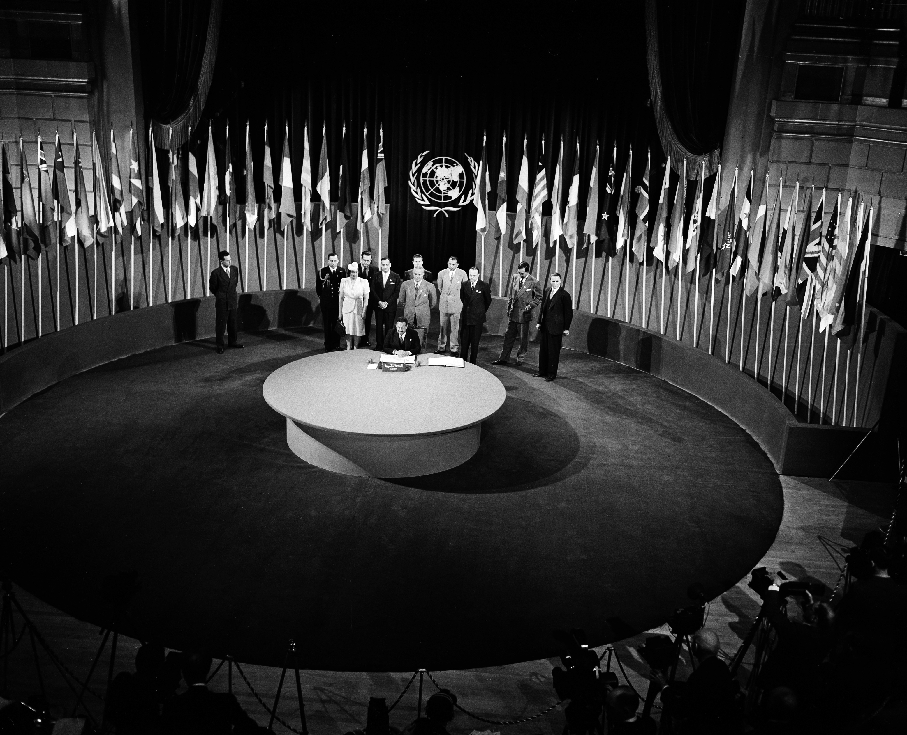 Black and white photo of a ceremonial event with a circular table and several flags.
