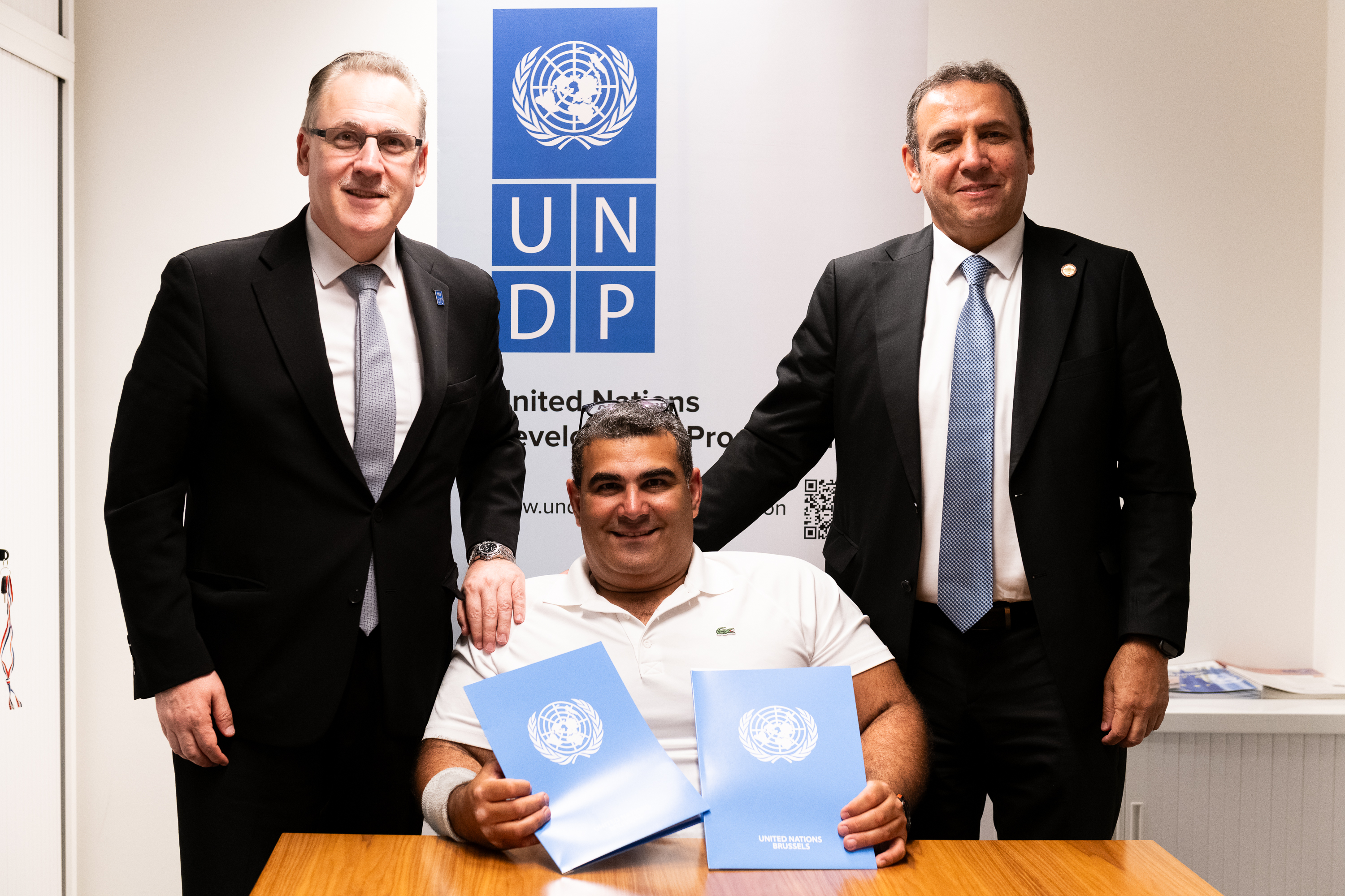 Three men pose together at a desk, holding documents, with a UNDP logo in the background.