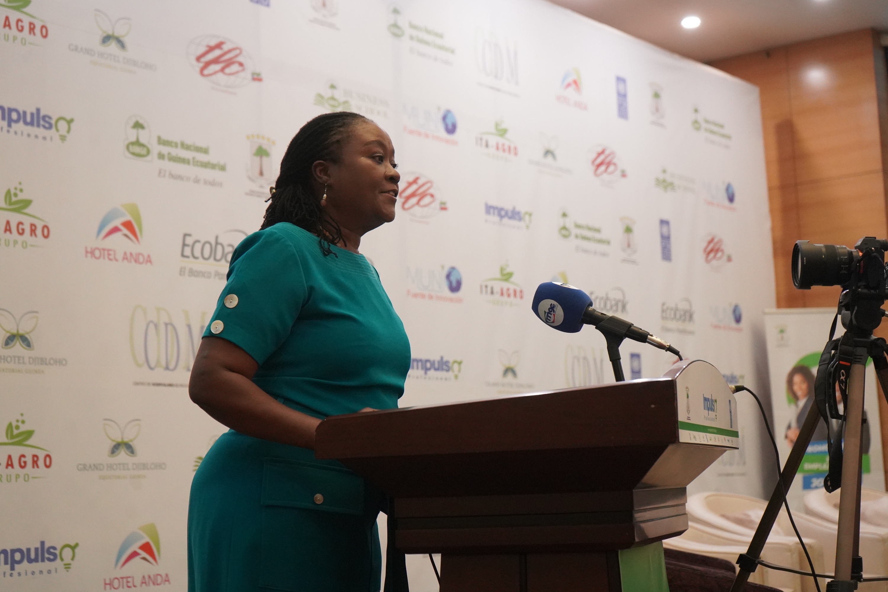 A woman in a teal dress speaks at a podium, with a camera and backdrop logos behind her.