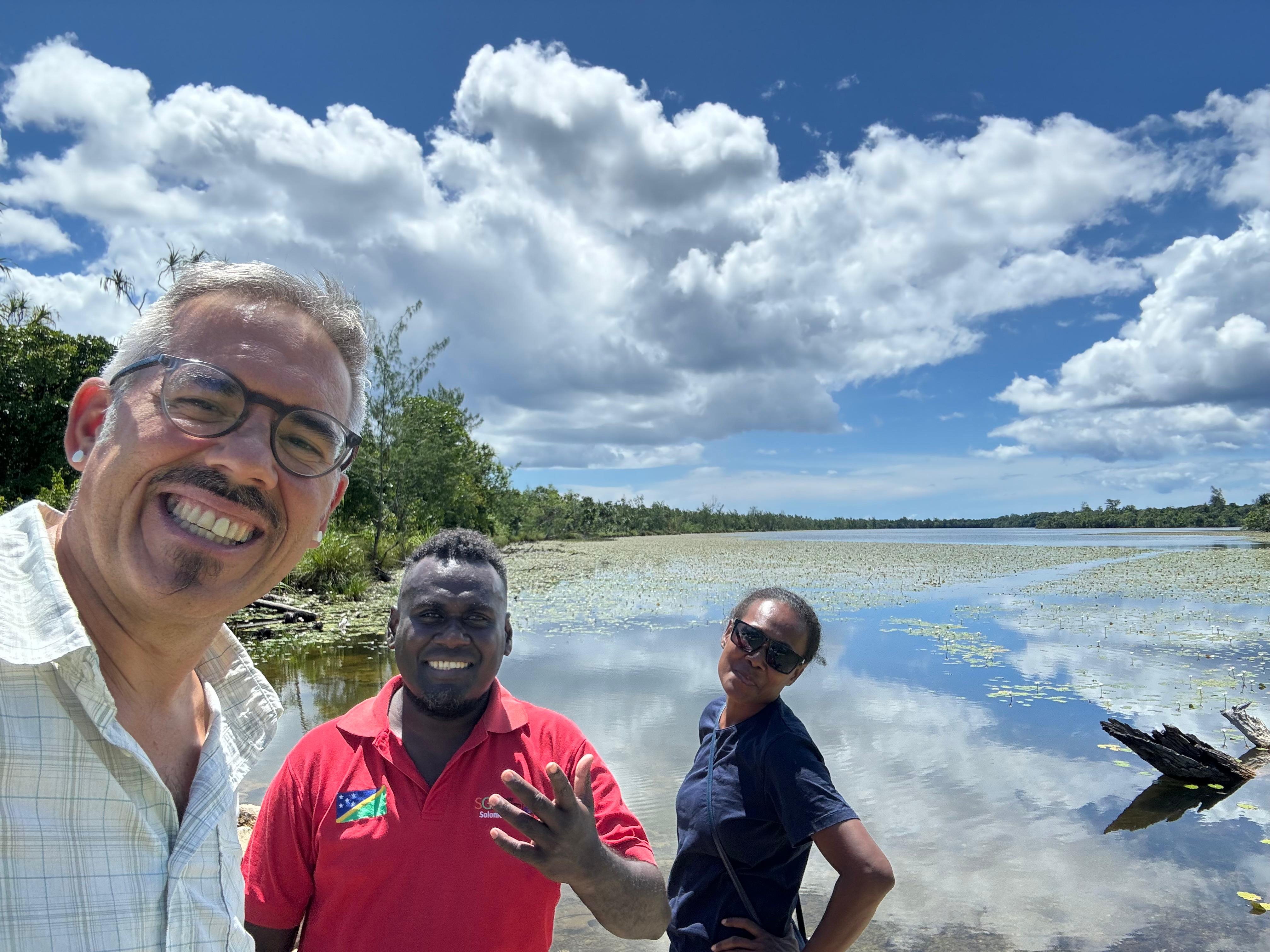 Three people smile together by a serene lake surrounded by greenery and blue skies.