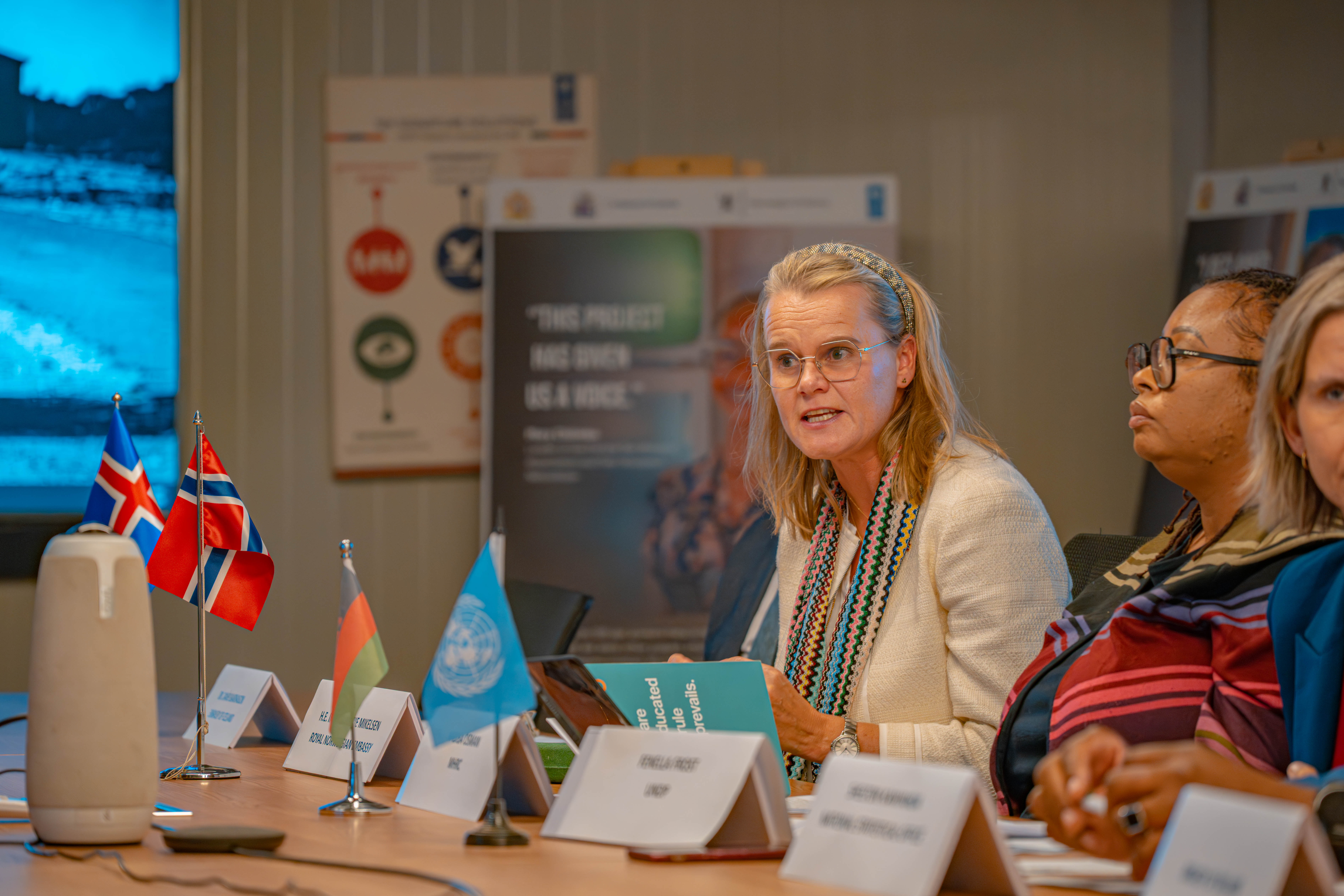 A woman with blonde hair speaks at a conference table, with flags and participants in the background.