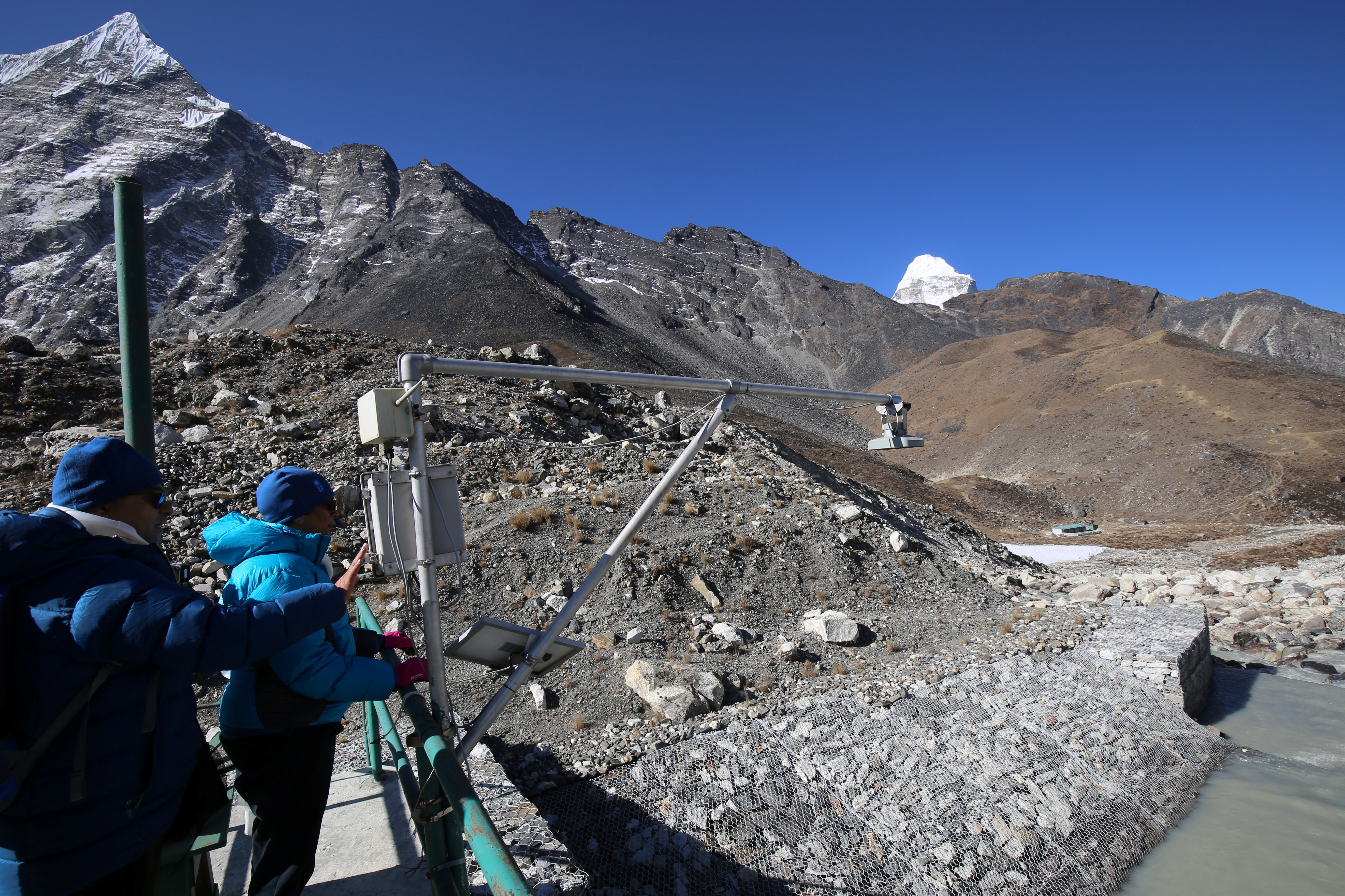 Two individuals in blue jackets observe a mountainous landscape with rocky terrain and a river.