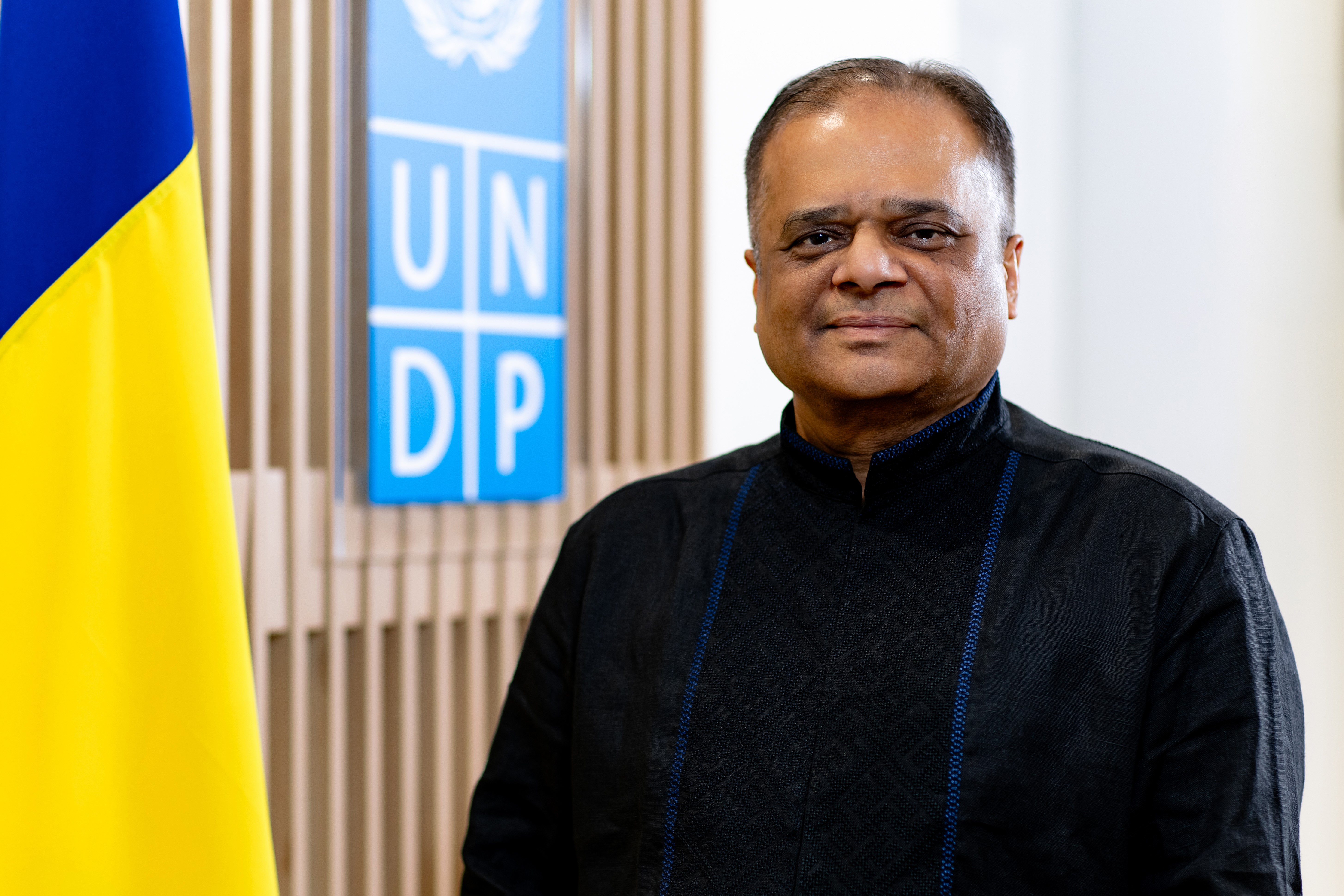 A man in traditional black attire stands in front of a UNDP banner and a yellow flag.