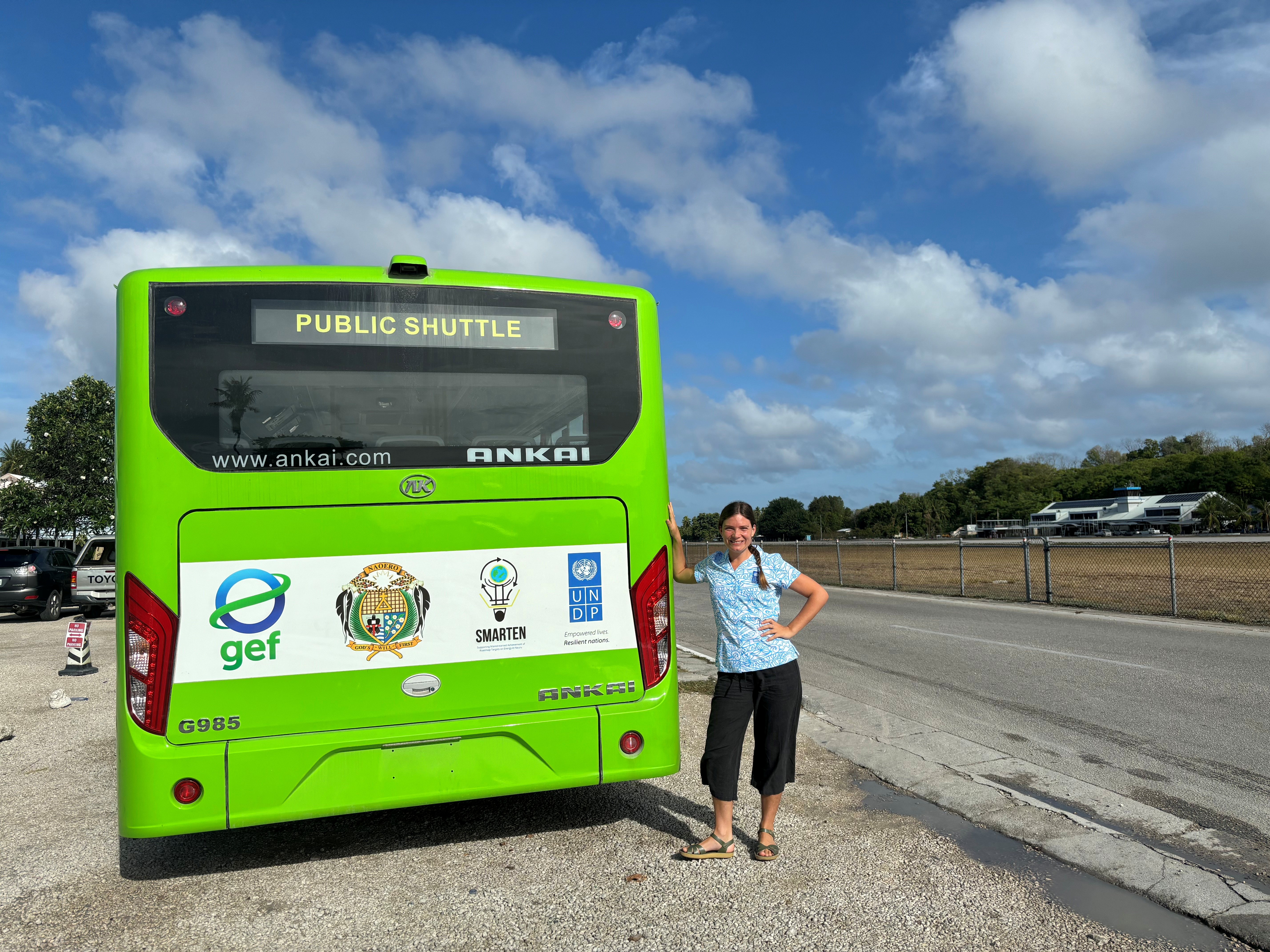 A woman stands beside a green public shuttle bus in a sunny outdoor setting.