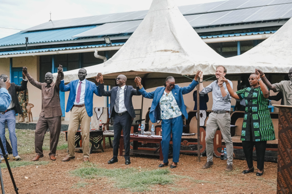 A group of people in formal attire holding hands and dancing outdoors, with tents in the background.