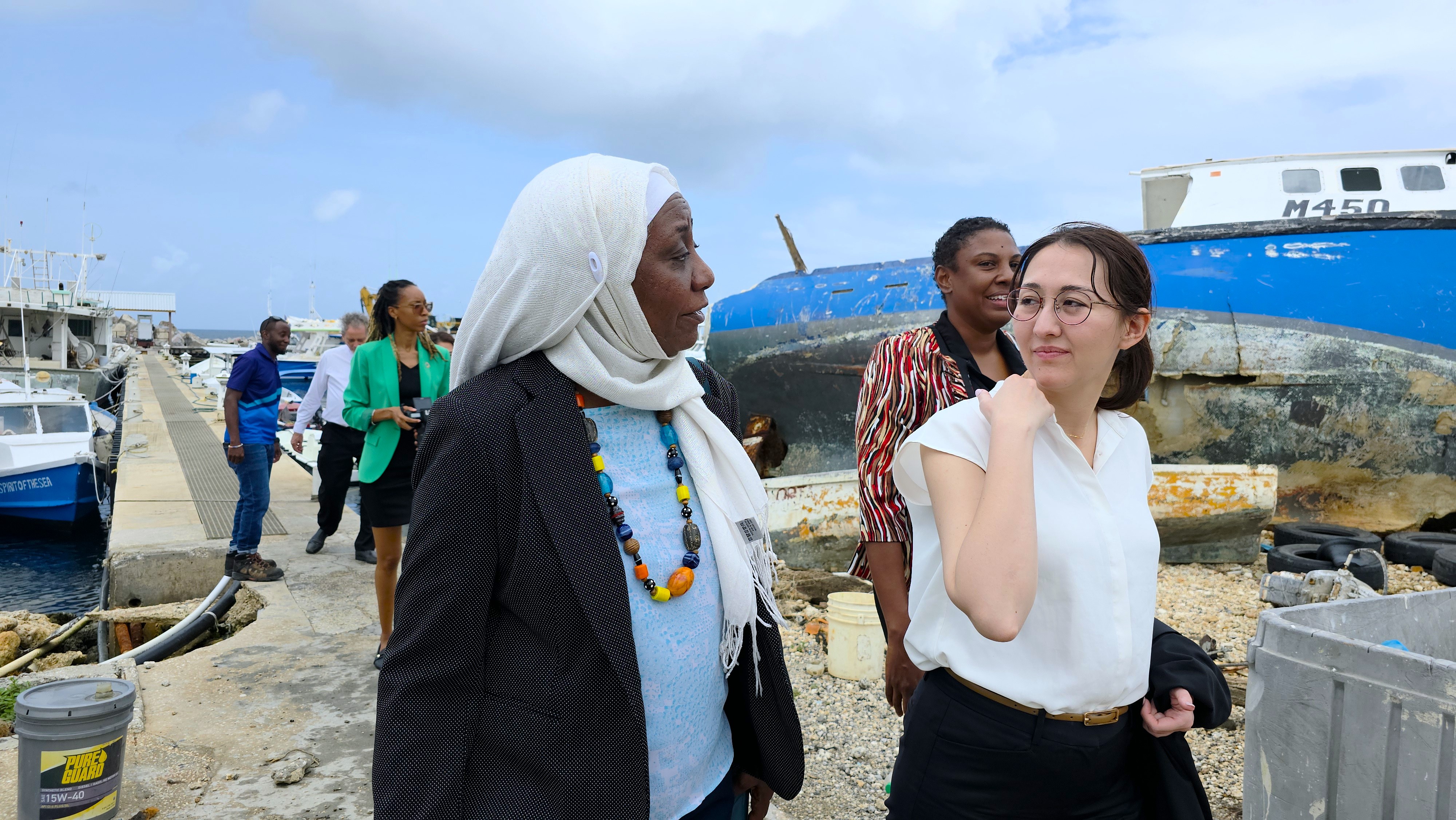 Two women walk along a harbor, engaged in conversation, with boats in the background.