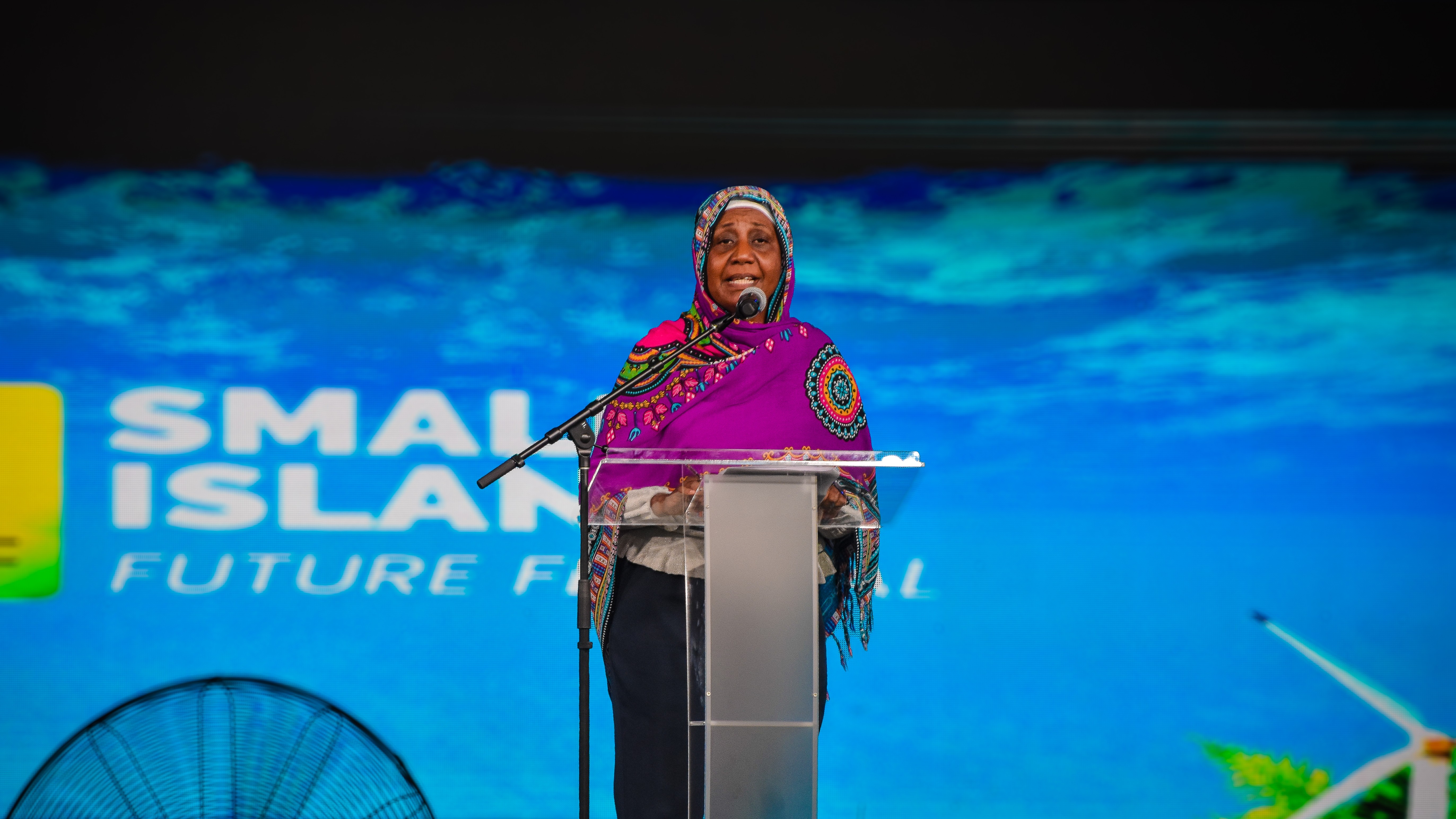 A woman in a colorful headscarf speaks at a podium, with a blue background and event branding.
