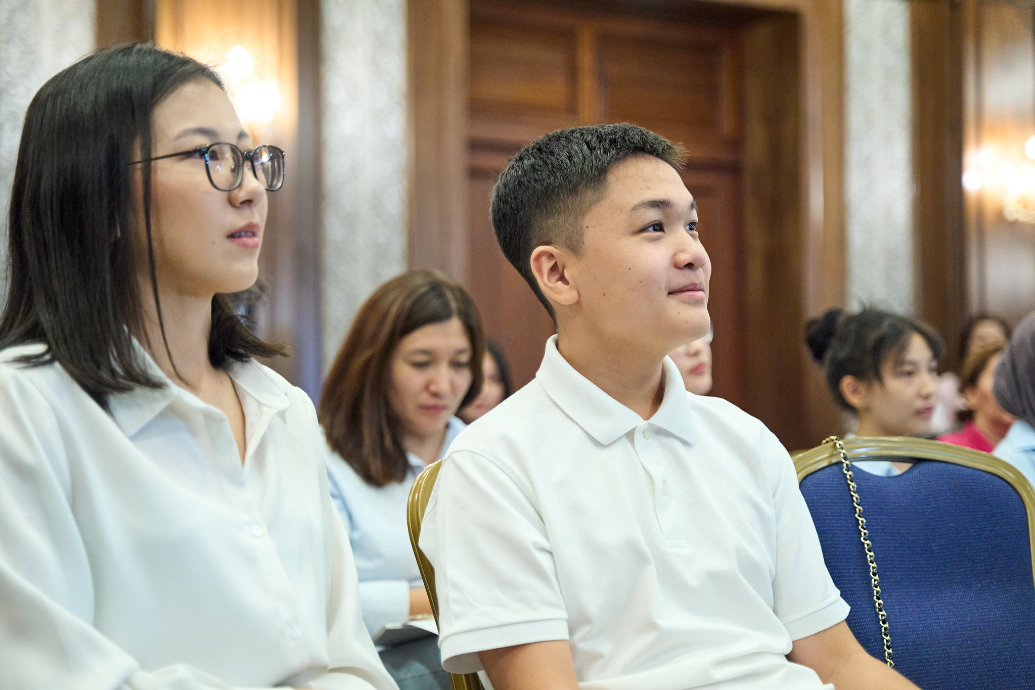 Two young individuals attentively listening during a presentation in a formal setting.
