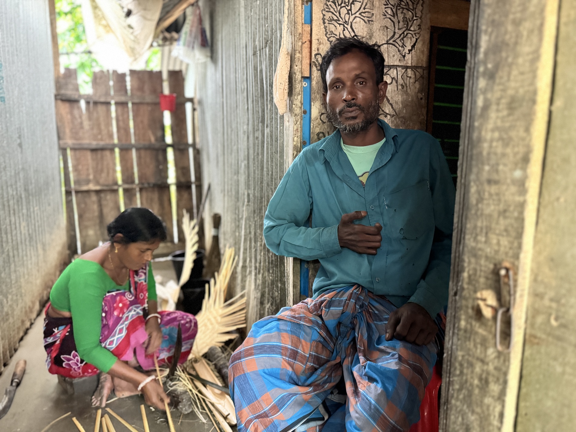 A man sits in a doorway while a woman weaves material behind him, both in traditional clothing.