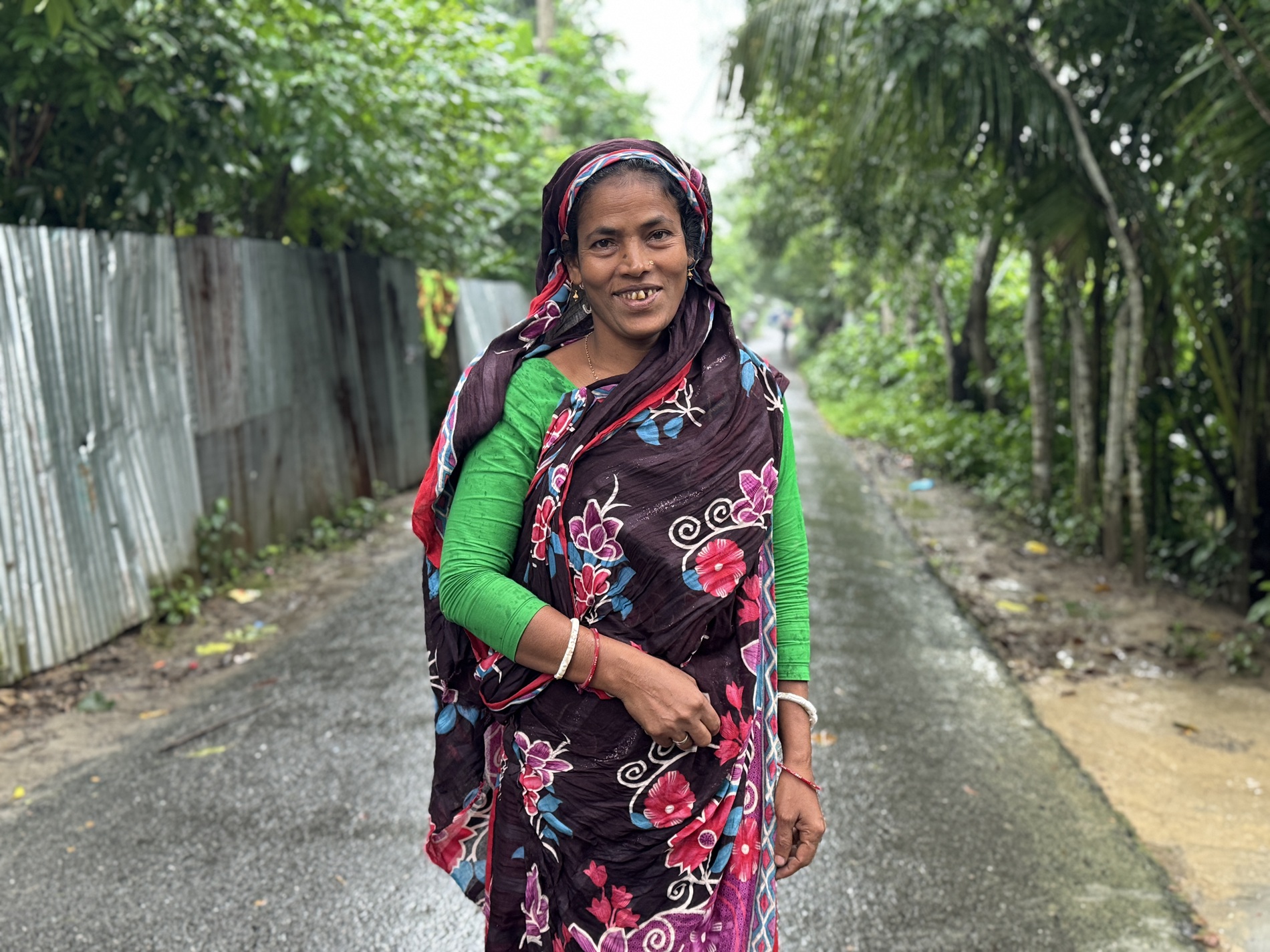 A woman in a colorful dress stands on a wet, tree-lined path, smiling warmly.
