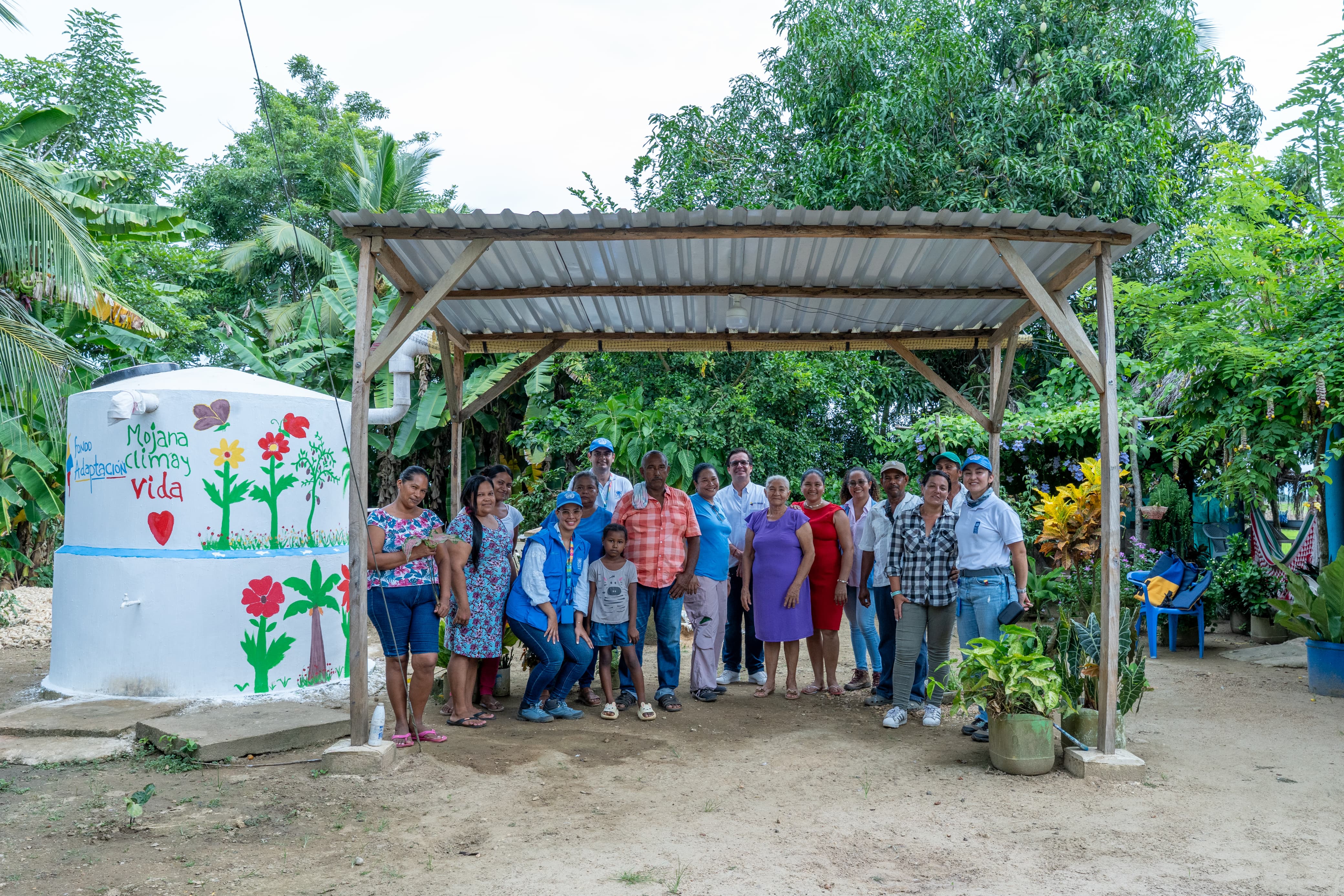 A group of people standing under a roofed structure, surrounded by greenery and colorful plants.