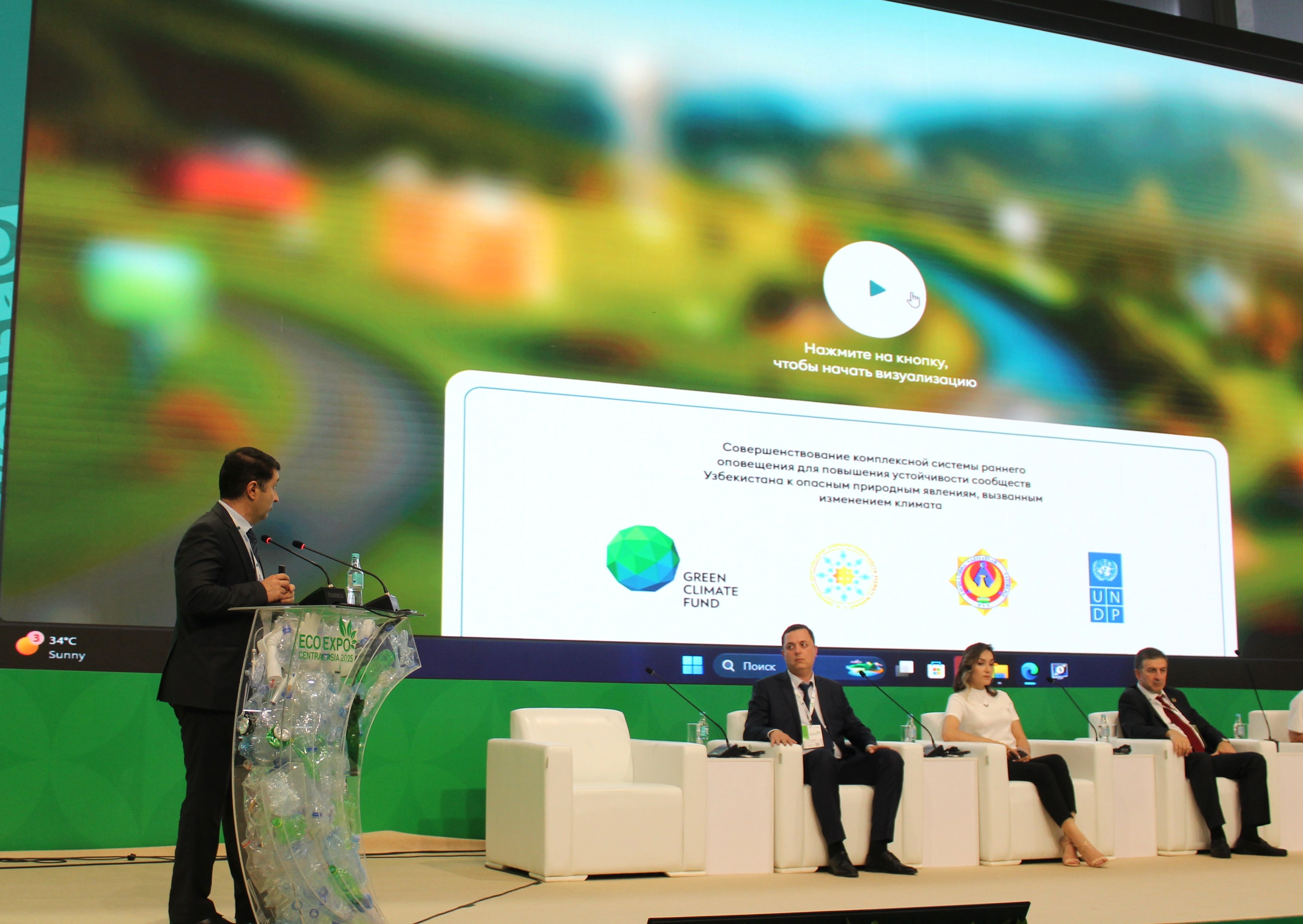 A speaker stands at a podium in front of a large screen displaying a landscape, with panelists seated.