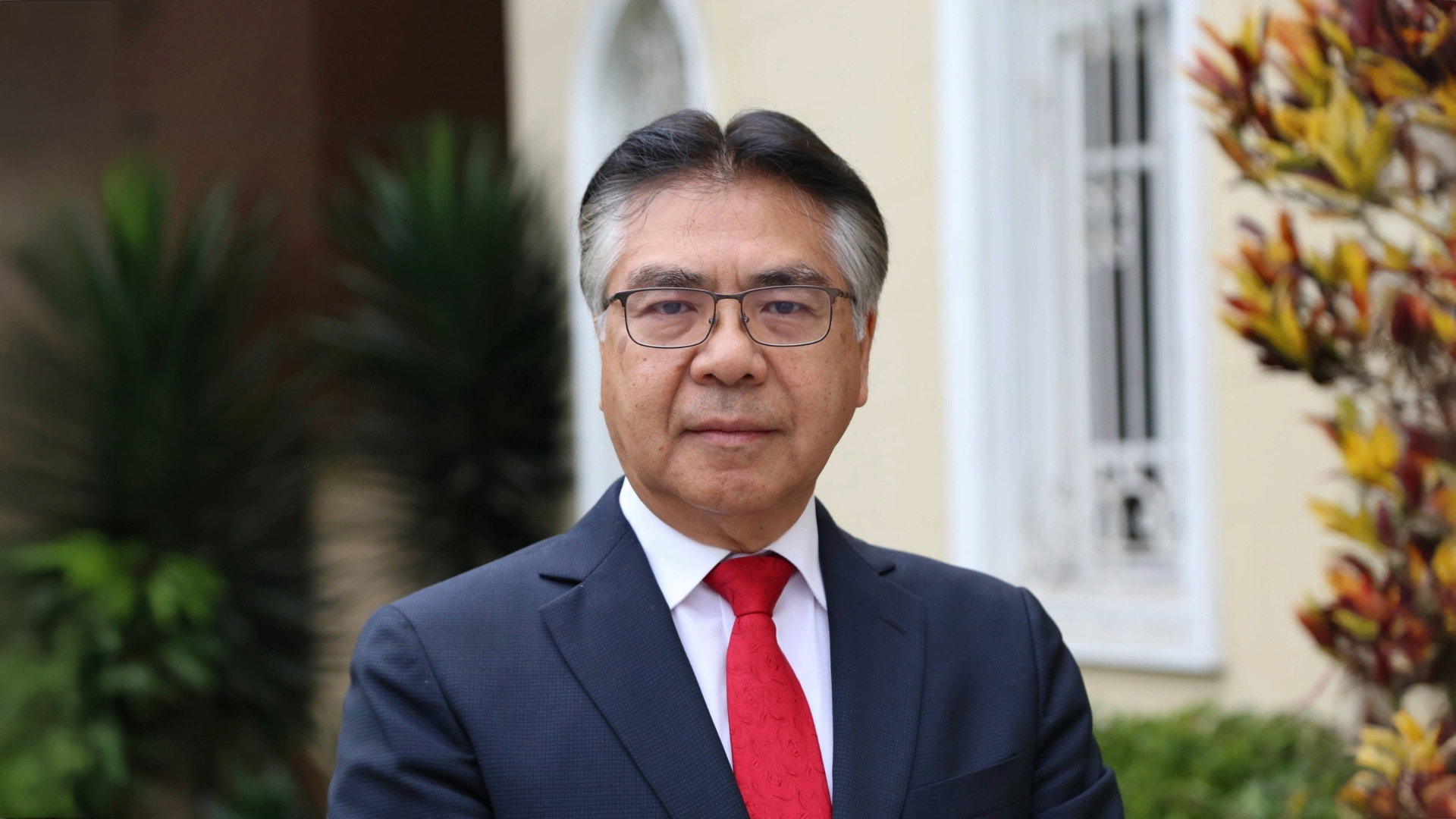 A man in a suit with a red tie stands outdoors, smiling slightly, with greenery in the background.
