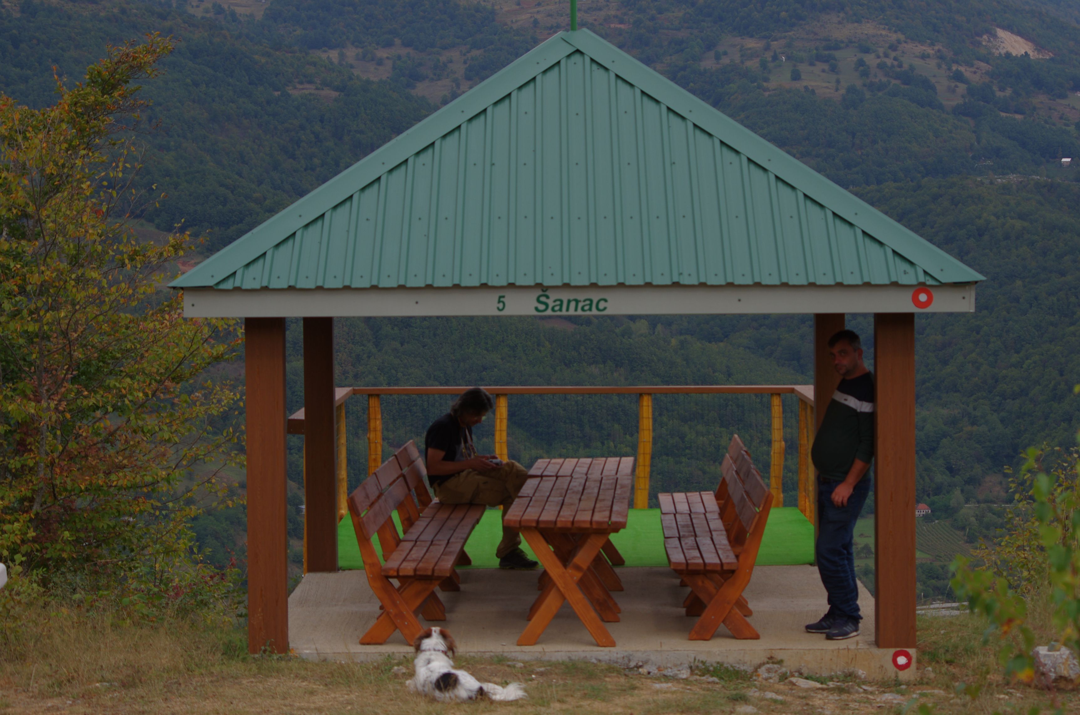 A covered wooden gazebo with two people and a dog, surrounded by mountains.