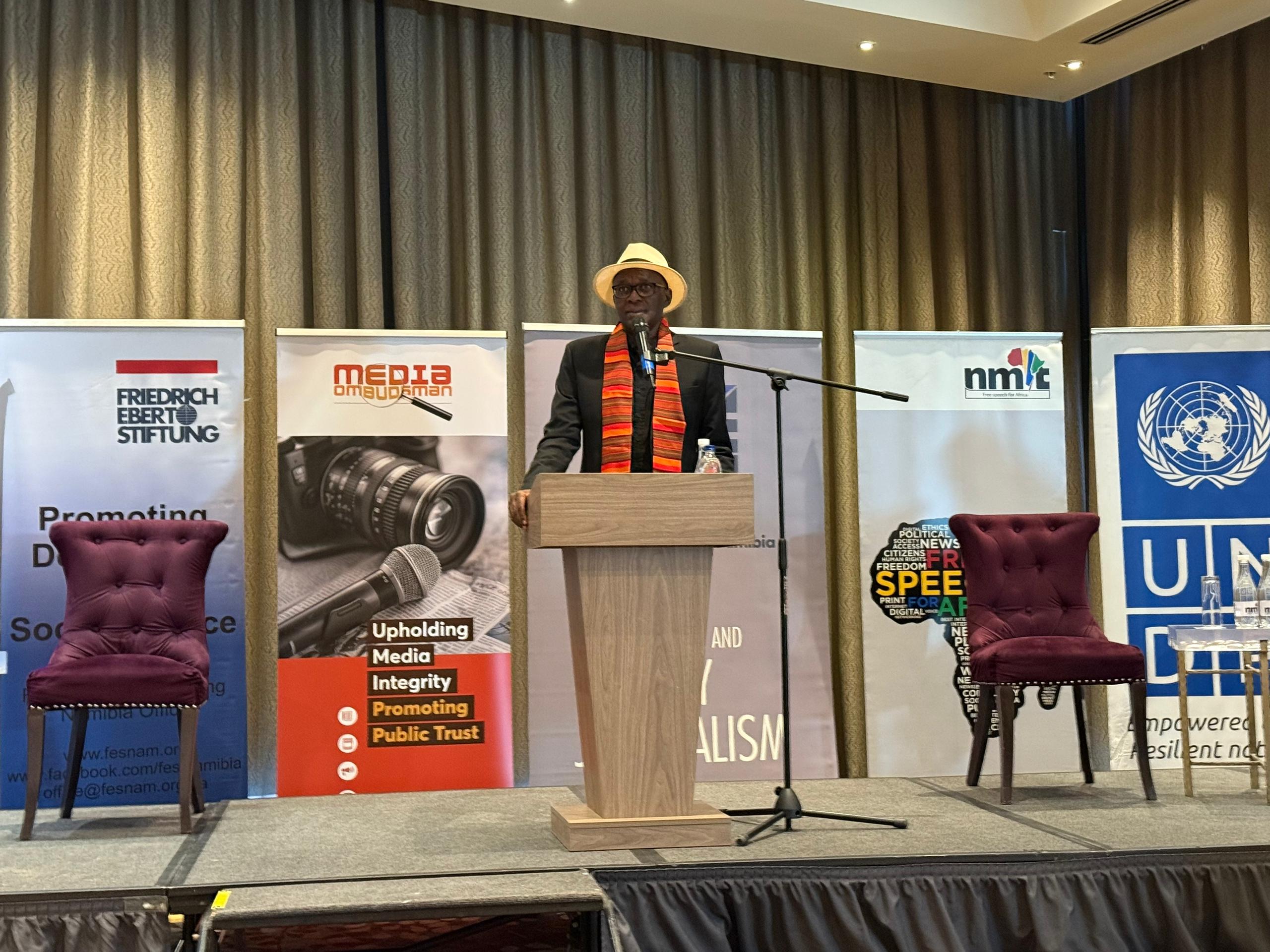 A speaker in a straw hat stands at a podium, addressing an audience in a conference setting.