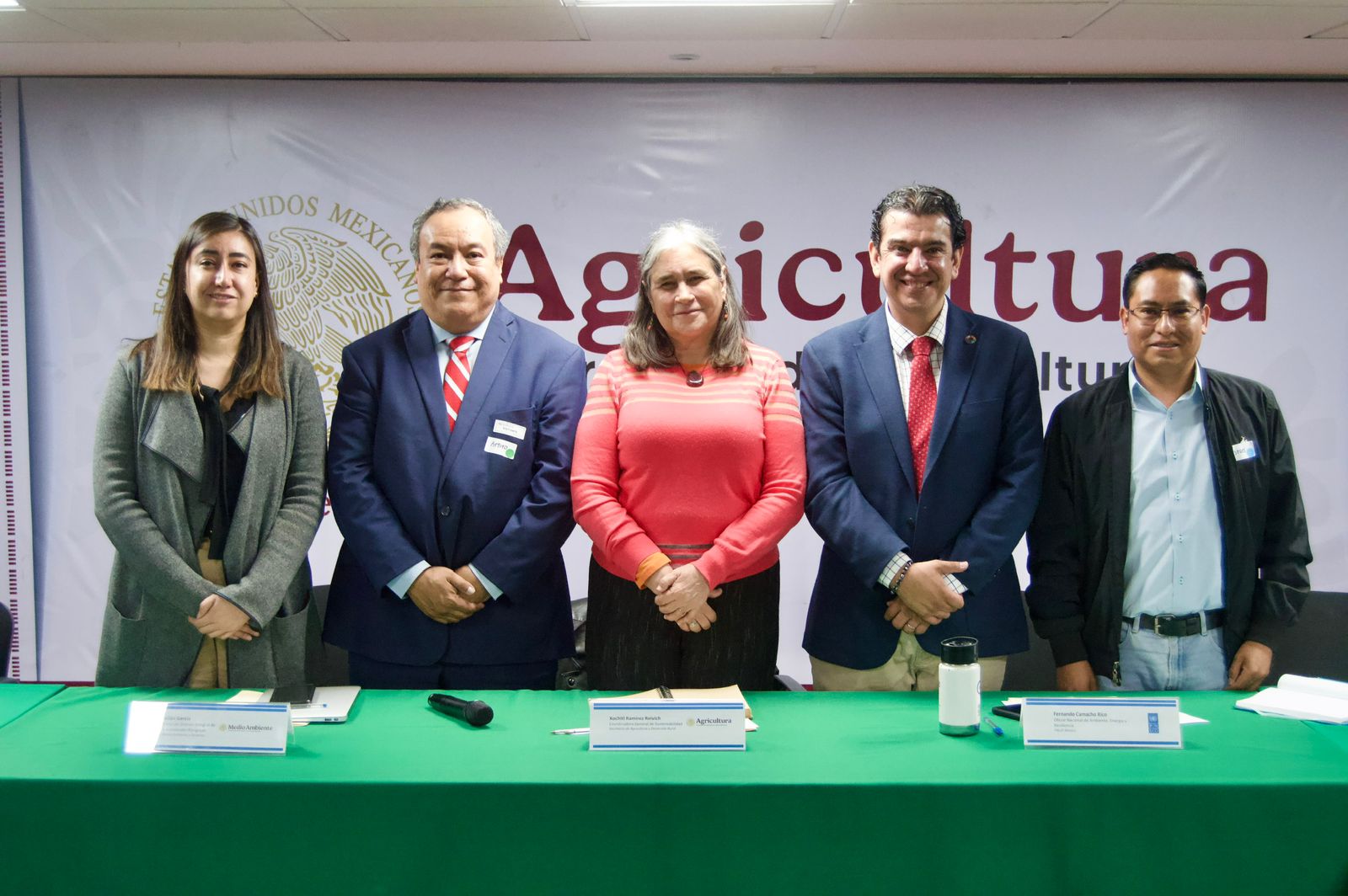 Five people stand behind a green table, smiling at the camera, in a conference setting.
