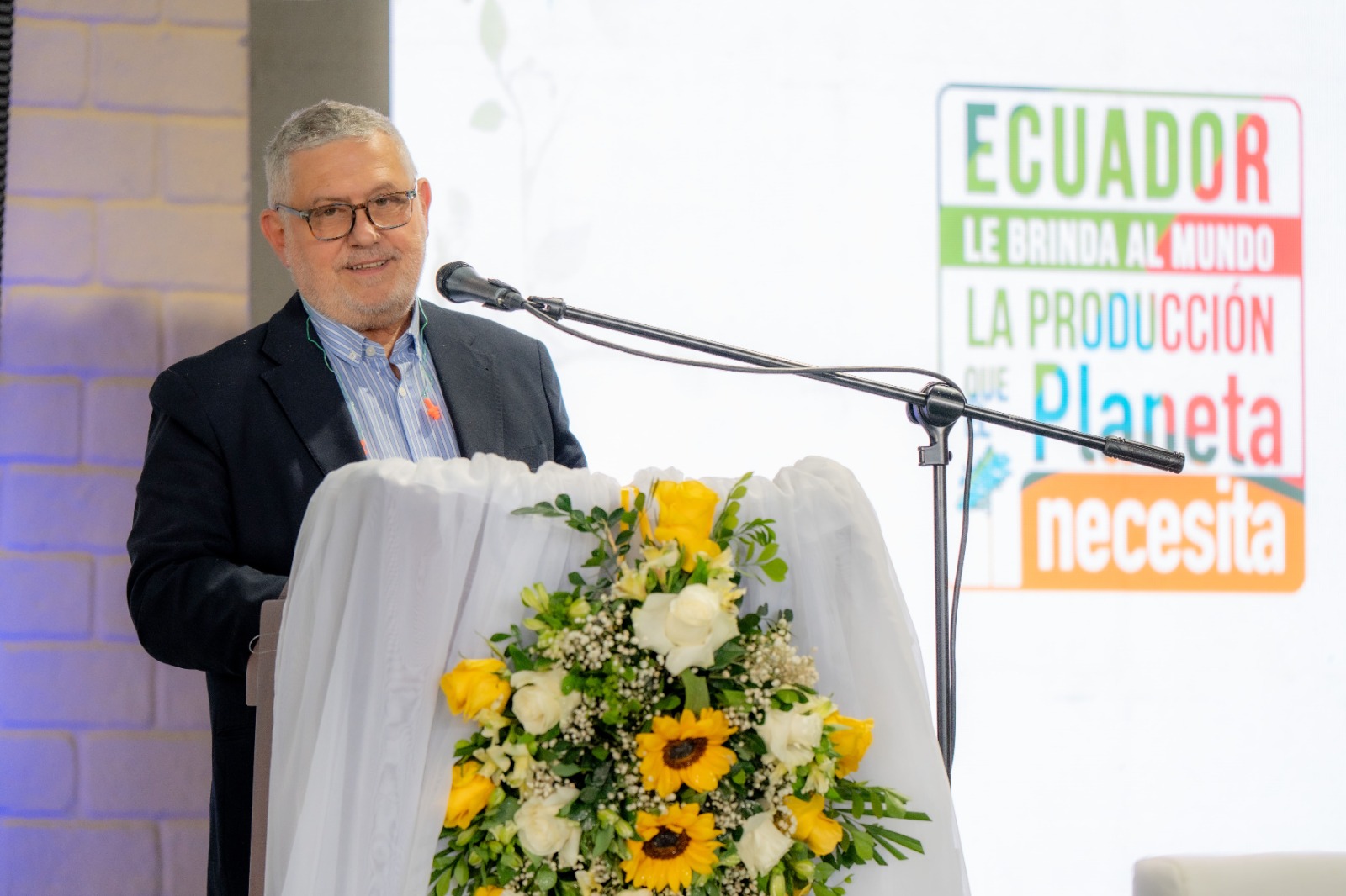 A man in a suit speaks at a podium adorned with flowers, displaying a colorful backdrop.