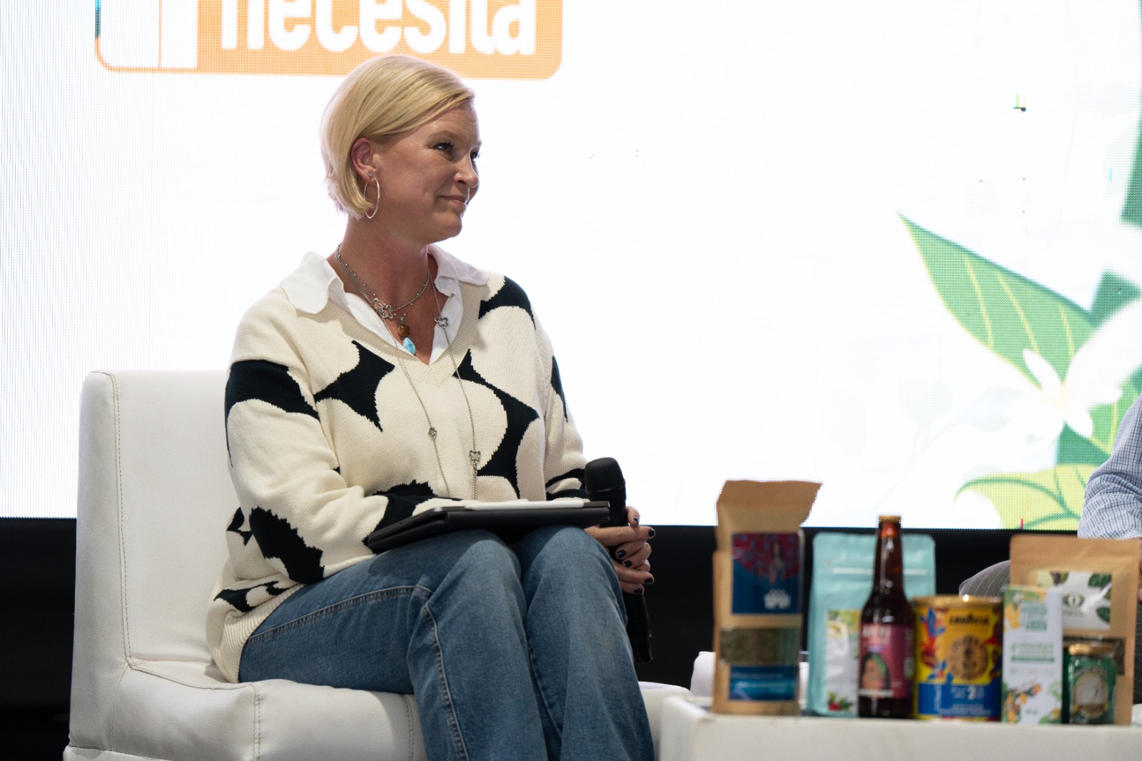 A woman in a black and white sweater sits on stage with products on a table in front of her.