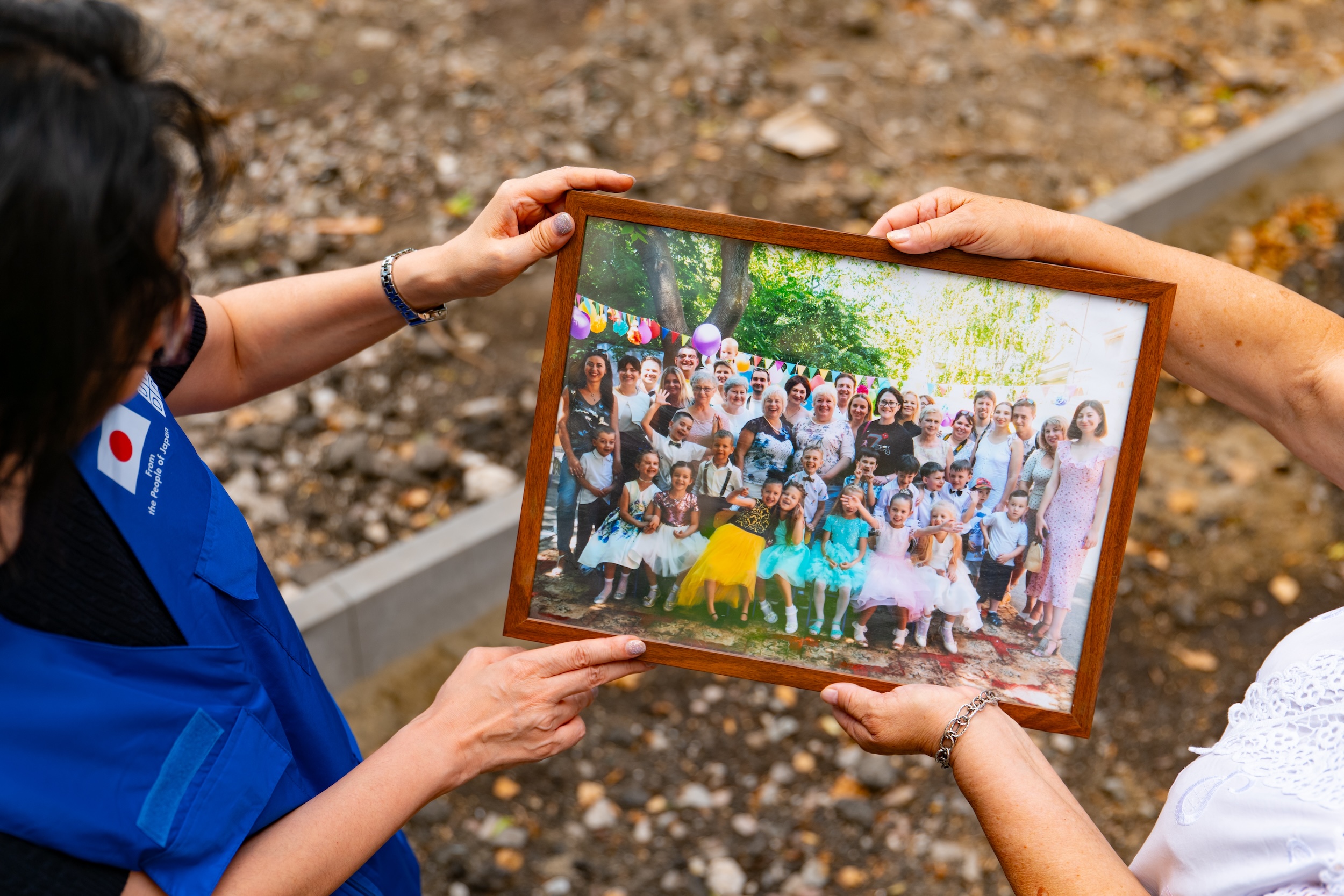 Two people hold a framed photo featuring a large group of children at an outdoor event.