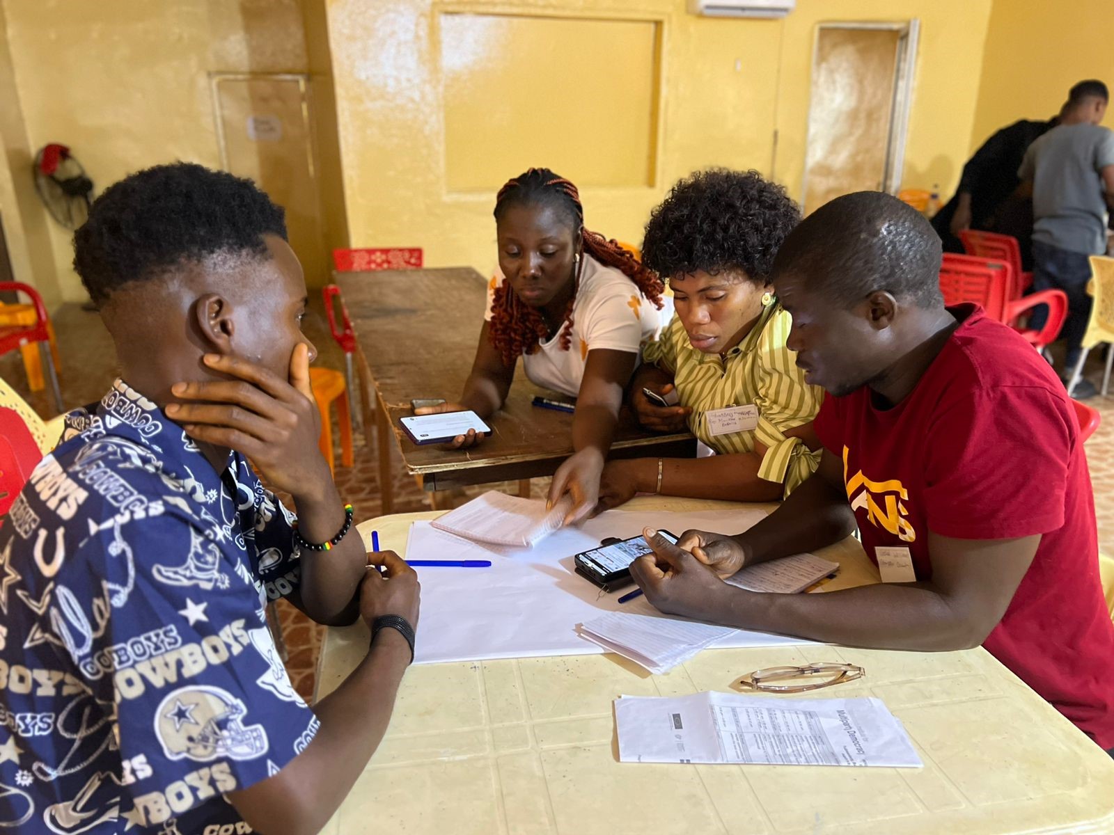 Four people collaborate around a table, discussing notes and using smartphones in a casual setting.