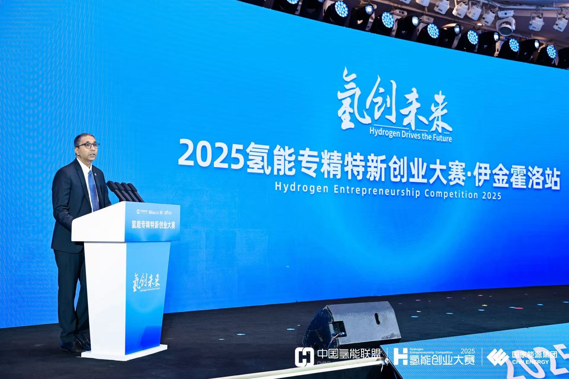 A speaker at a podium presents at a Hydrogen Entrepreneurship Competition with a blue backdrop.