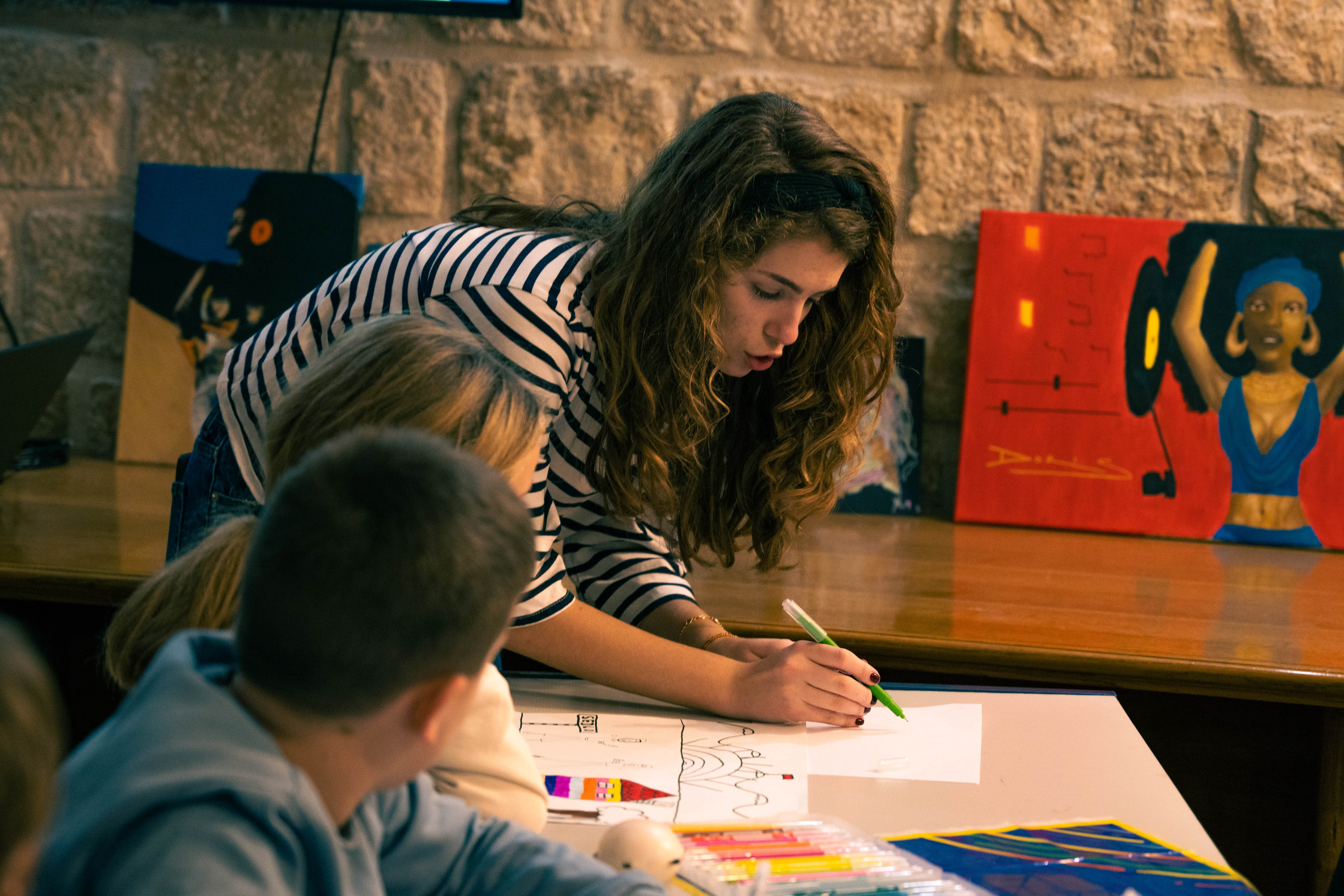 A young woman with curly hair draws on a paper while children observe attentively.